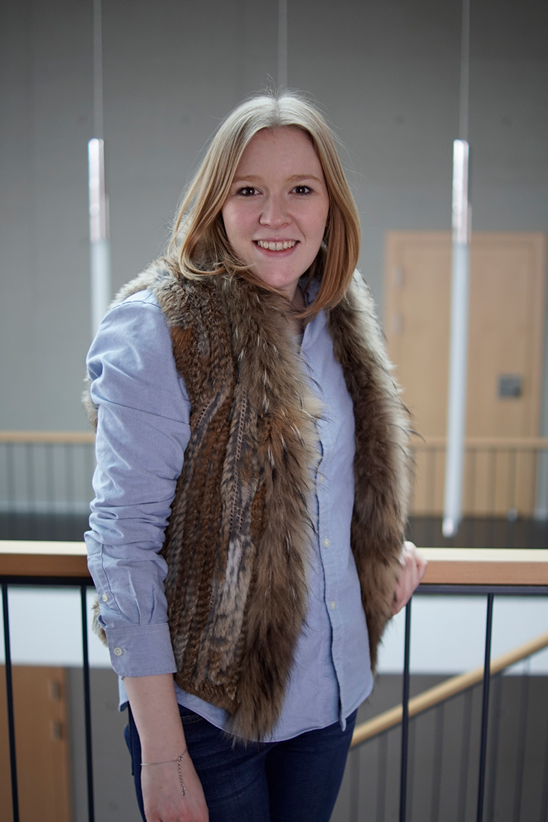 Eine Person mit langen Haaren lehnt lächelnd an einem Geländer und trägt ein blaues Hemd und eine Pelzweste. Der Hintergrund erinnert an das Designethos der Hochschule Coburg und zeigt ein minimalistisches Interieur, das durch vertikale Leuchten und Holzakzente bereichert wird.