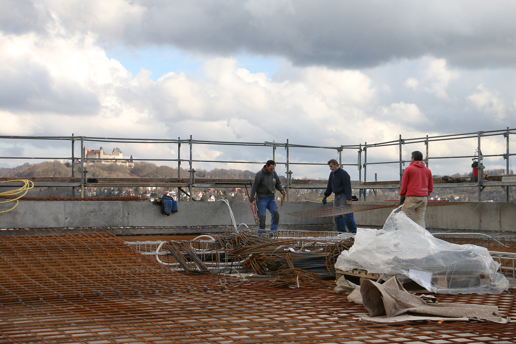 Drei Bauarbeiter stehen auf einer Baustelle mit Blick auf eine malerische Landschaft. In der Ferne sind die Hochschule Coburg und ein Schloss zu sehen. Auf der Baustelle sind Metallgitter, Werkzeuge und Geräte verstreut, während ein wolkiger Himmel den Hintergrund bildet.