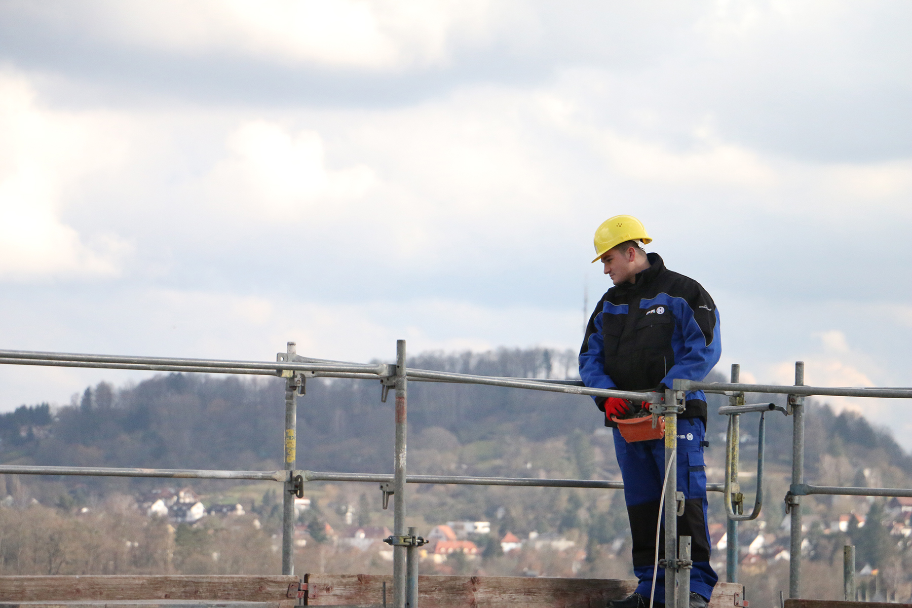 Ein Bauarbeiter in blau-schwarzer Uniform und gelbem Bauhelm steht stolz auf einem Gerüst der Hochschule Coburg und hält sich am Geländer fest. Im Hintergrund erstreckt sich eine weite Landschaft und ein wolkiger Himmel.