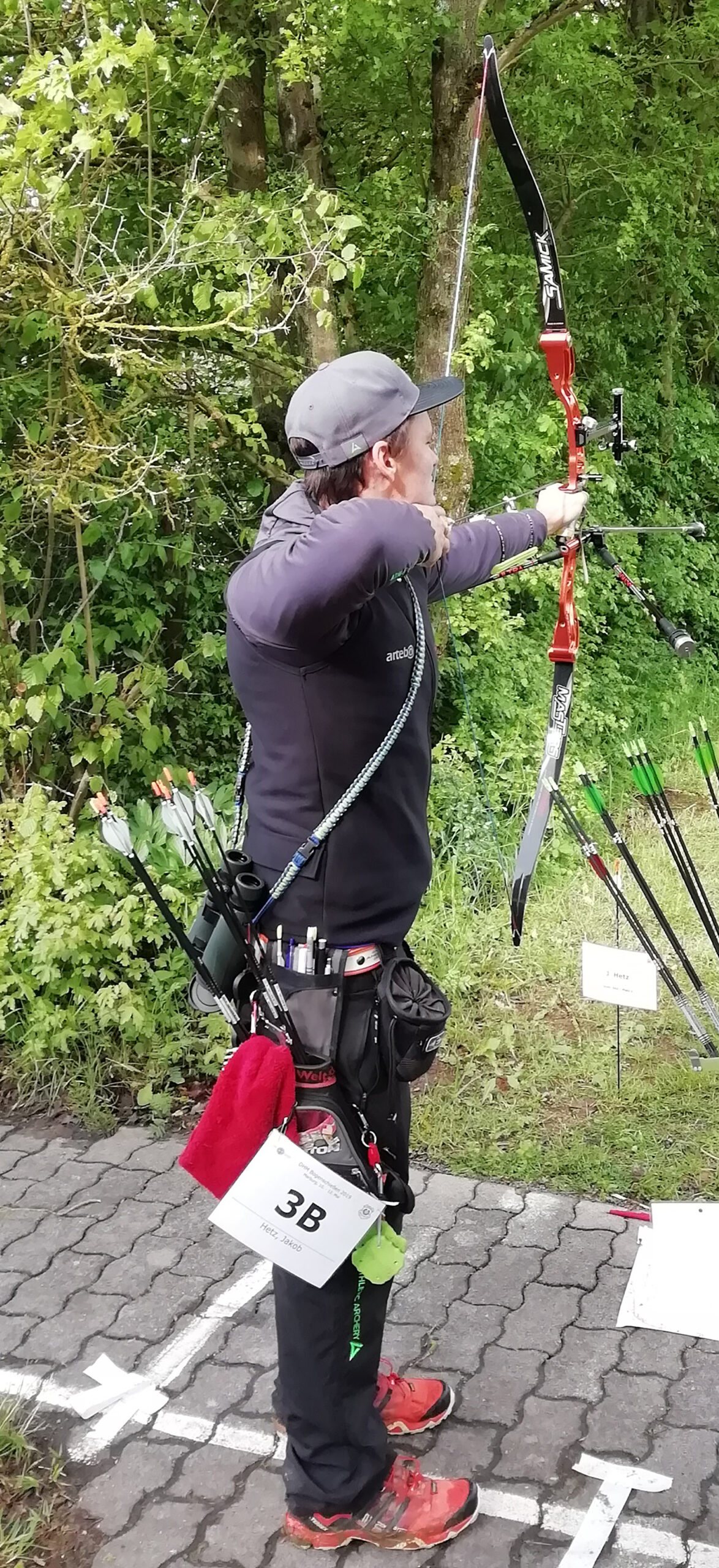 Eine Person in Outdoor-Ausrüstung steht auf einer gepflasterten Fläche der Hochschule Coburg und richtet einen rot-schwarzen Recurvebogen auf ein Ziel. Sie ist von Grün umgeben, trägt eine Mütze und hat Pfeile in einem Köcher. An ihrer Hüfte ist eine nummerierte Karte mit der Aufschrift „3B“ befestigt.