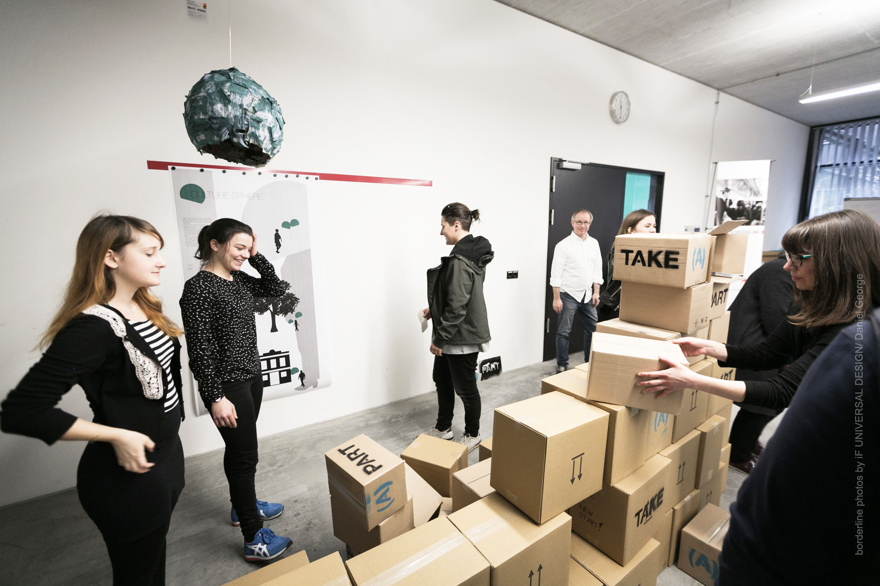 Eine Gruppe von Menschen beschäftigt sich in einem Büroraum mit großen Fenstern. Zahlreiche Kartons mit den Aufschriften „PAR“ und „TAKE“ sind in der Nähe gestapelt und deuten auf ein laufendes Projekt der Hochschule Coburg hin. Ein dekorativer Globus, der von der Decke hängt, verleiht der akademischen Atmosphäre einen Hauch von Verspieltheit.