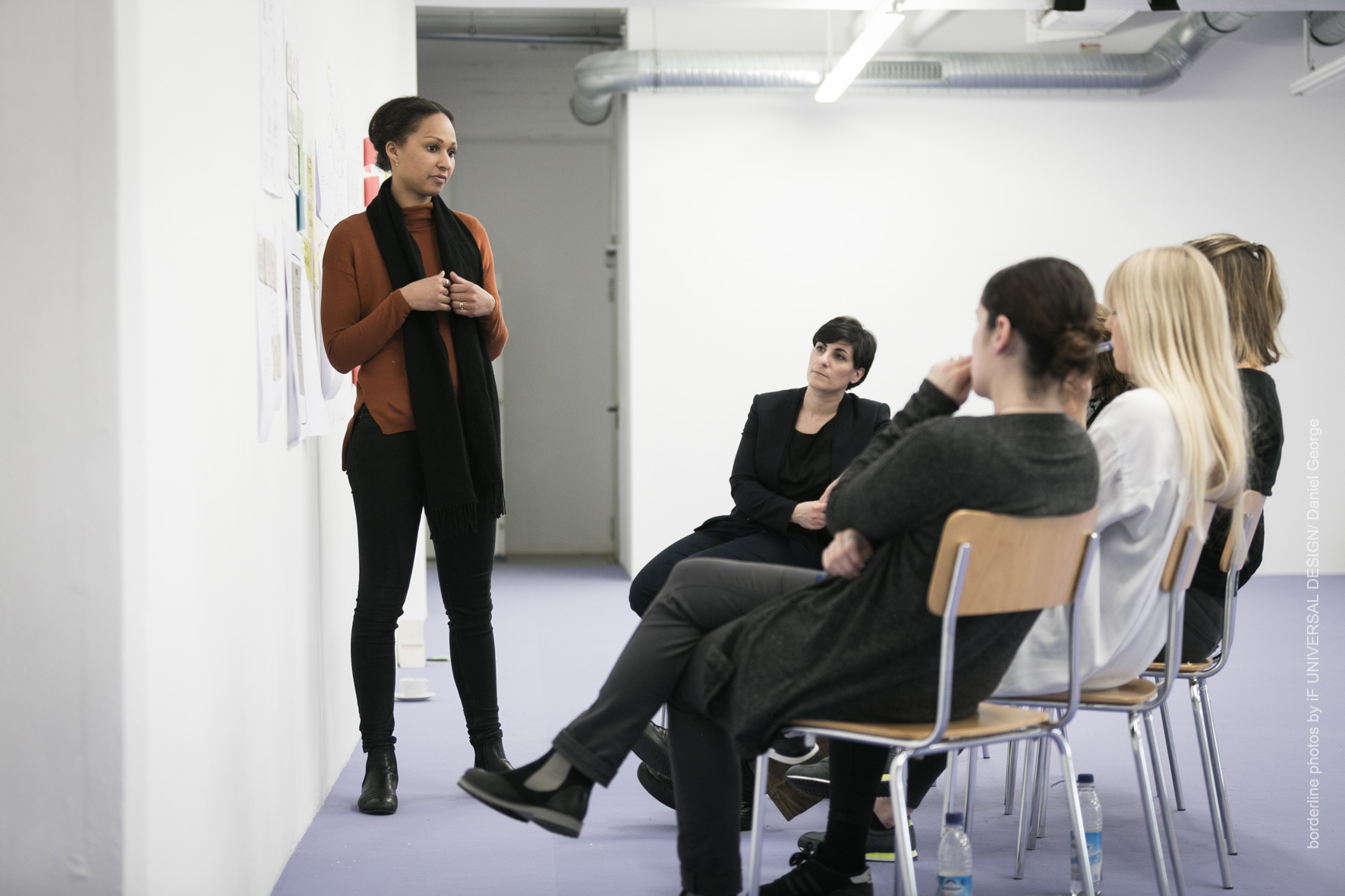 Eine Frau steht vor einer Gruppe und hält einen Vortrag in einem modernen, minimalistischen Raum der Hochschule Coburg. Vier Personen sitzen und hören aufmerksam zu. An den Wänden hängen mehrere Blätter Papier, was die professionelle und konzentrierte Atmosphäre verstärkt.