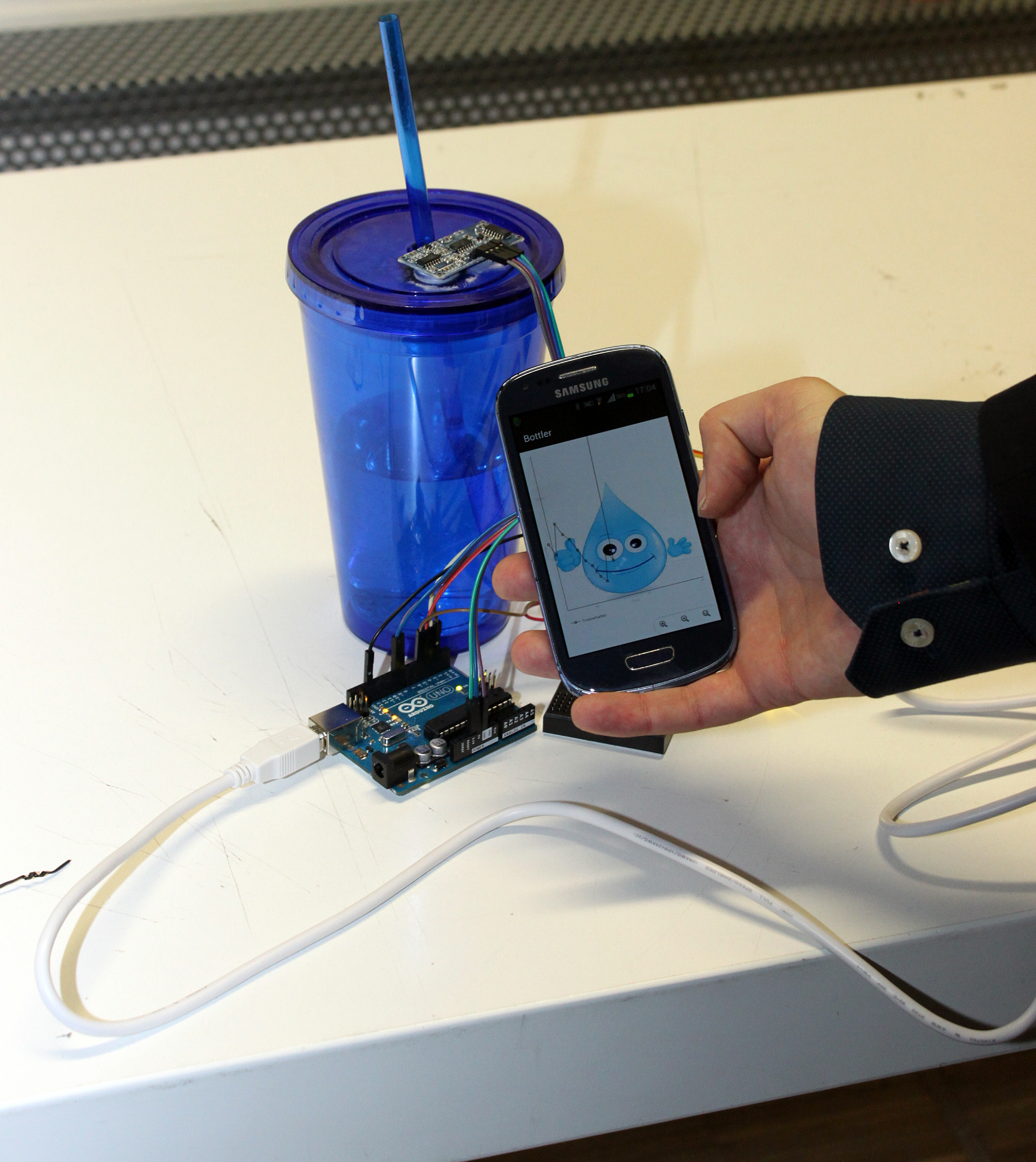 Eine Person der Hochschule Coburg hält ein Smartphone, auf dem eine App mit einem Wassertropfen-Charakter angezeigt wird. Das Telefon ist mit einem Arduino-Board verbunden, das wiederum mit einem blauen Becher mit Strohhalm auf dem Tisch verkabelt ist.