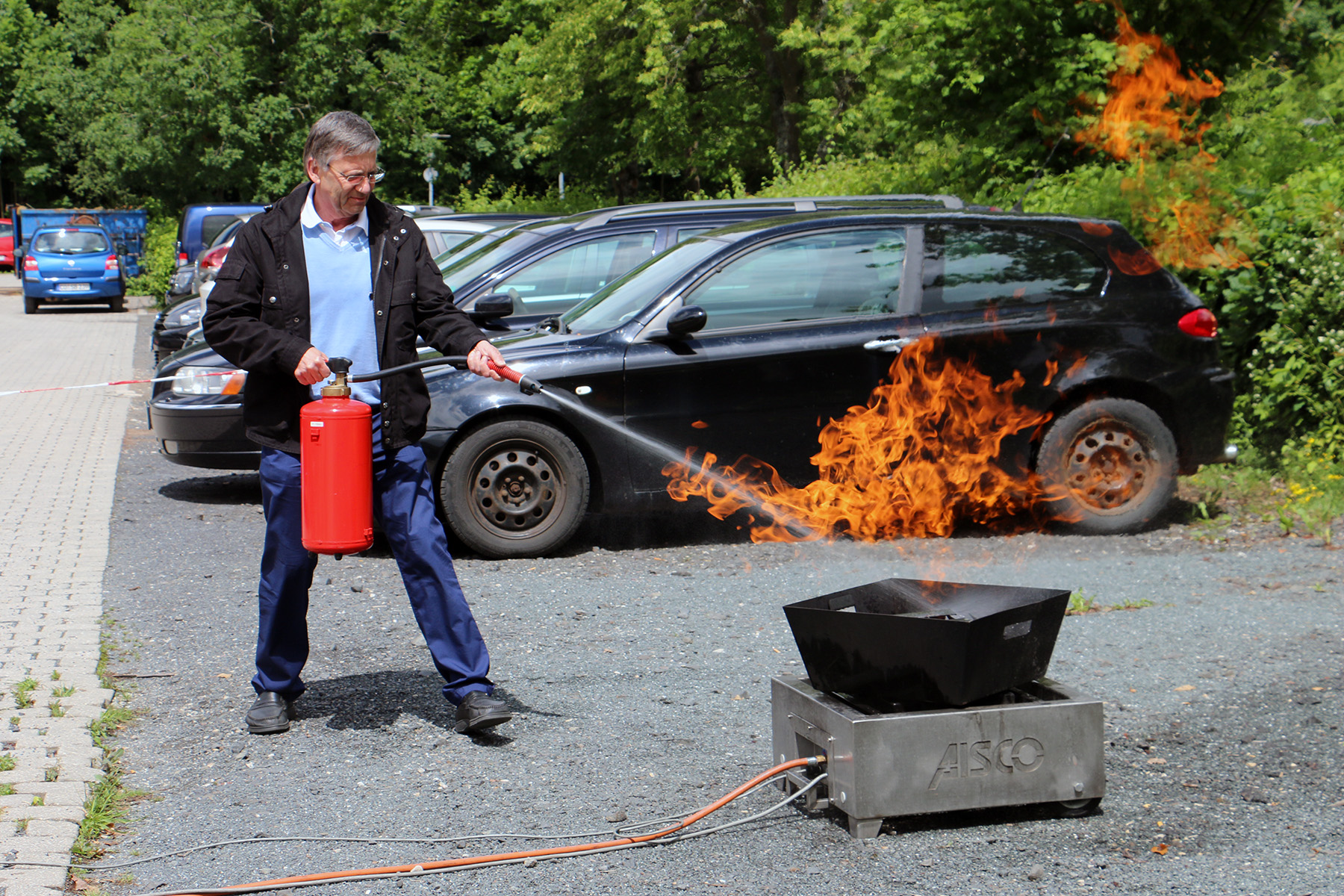 Ein Mann löscht mit einem Feuerlöscher Flammen aus einem Metallcontainer im Außenbereich nahe der Hochschule Coburg. Im Hintergrund vervollständigen mehrere geparkte Autos und Bäume die Szene.