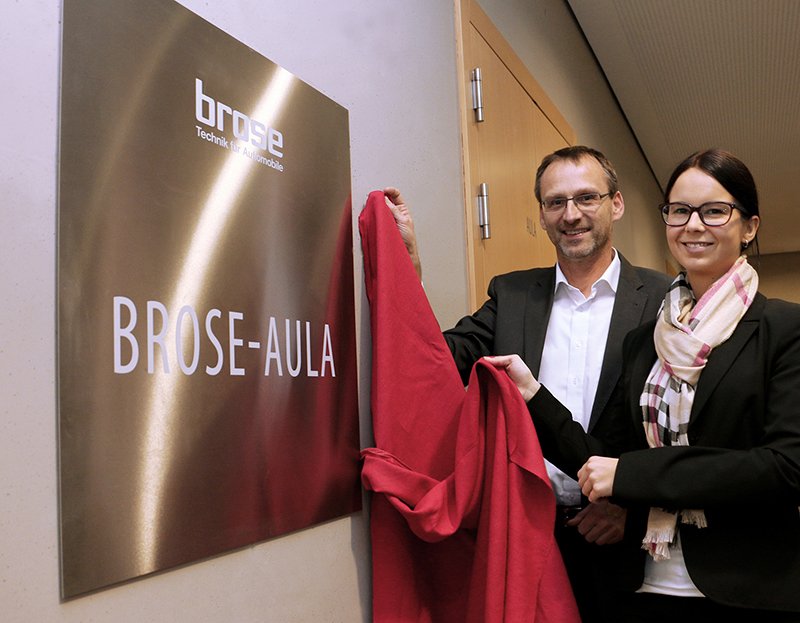 Zwei Personen enthüllen ein Schild mit der Aufschrift „Brose-Aula“ an einer Wand der Hochschule Coburg. Der Mann trägt einen Anzug, die Frau einen Blazer und einen Schal. Beide lächeln, während sie den roten Stoff halten, der für die Enthüllung verwendet wurde, ein stolzer Moment für die Institution.