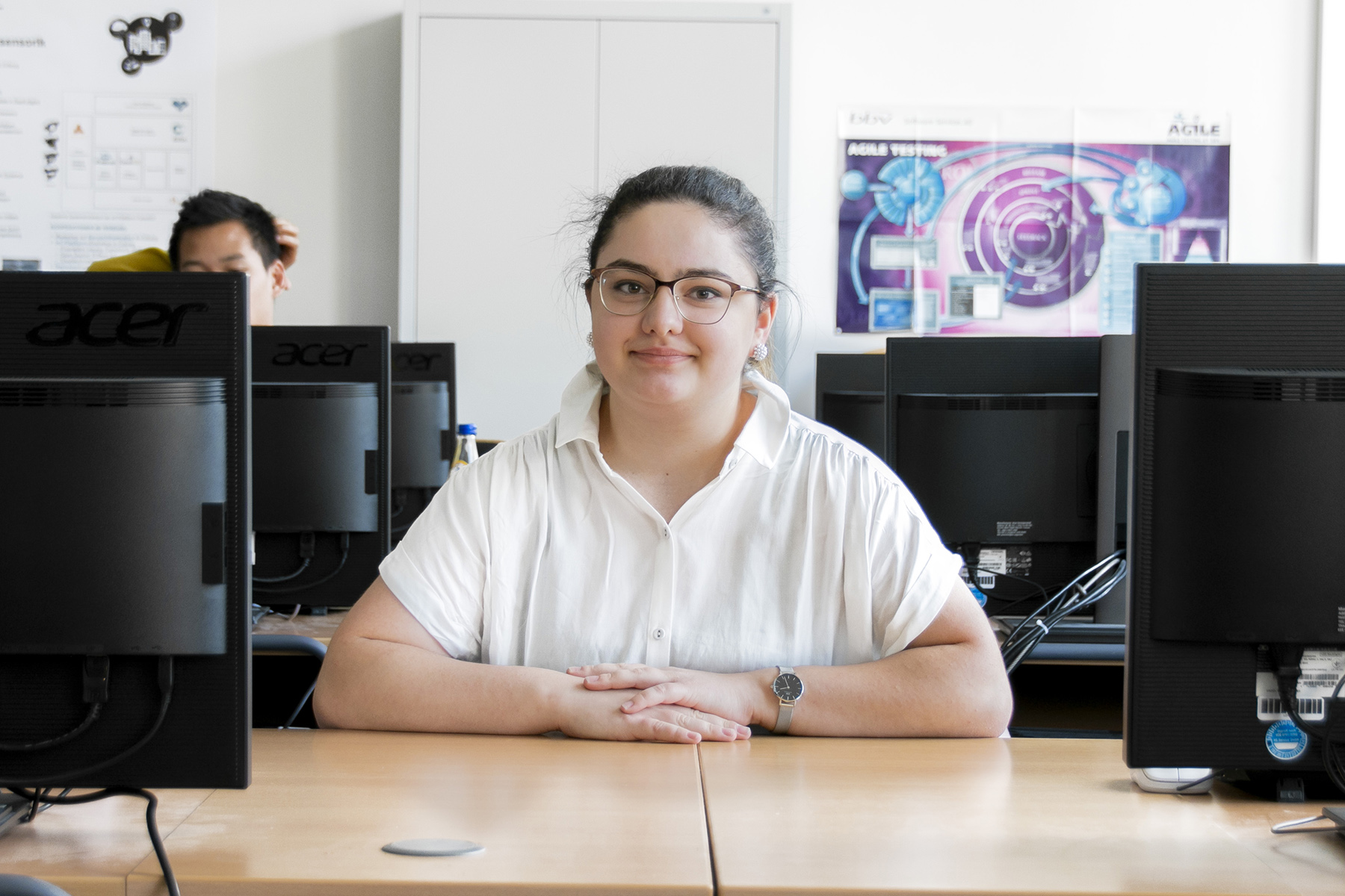 Eine Person mit Brille und weißem Hemd sitzt an einem Schreibtisch mit Computern in einem Büro oder einem Unterrichtsraum der Hochschule Coburg. Im Hintergrund ist eine weitere Person teilweise zu sehen. Ein lila Poster bringt Farbe an die Wand.