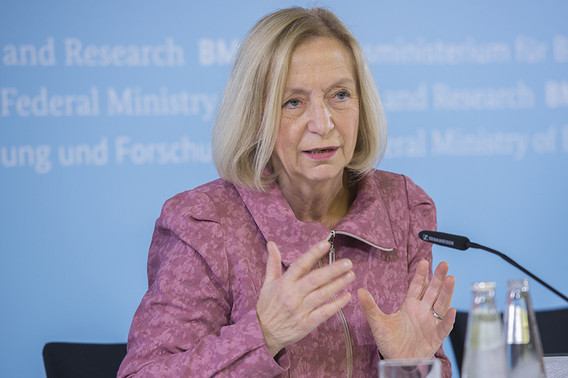 Eine Person mit blondem Haar spricht auf einer Pressekonferenz der Hochschule Coburg und gestikuliert lebhaft in einer rosa Jacke. Der verschwommene Text auf der blauen Wand weist auf das Thema der Veranstaltung hin. Zwei Mikrofone und eine Wasserflasche stehen prominent auf dem Tisch, während sie das Publikum anspricht.