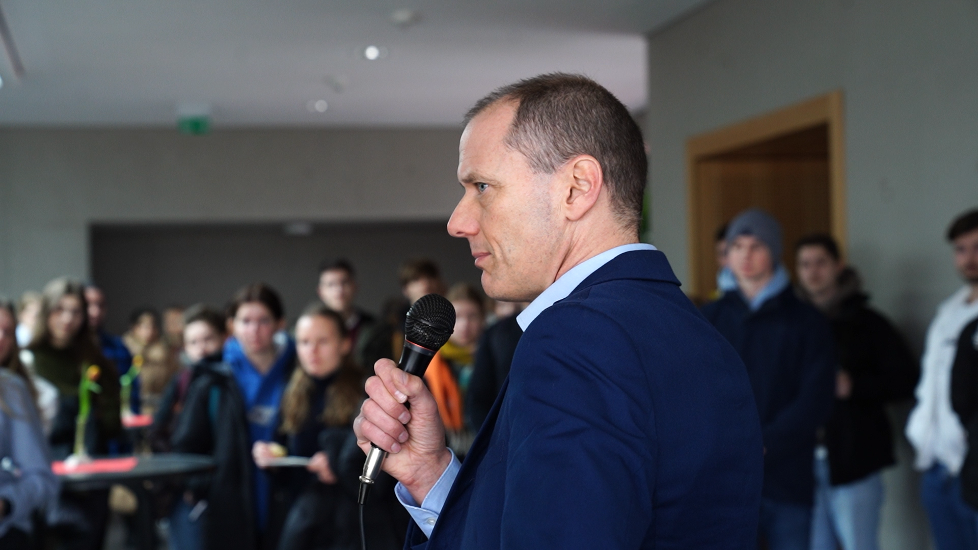 Ein Mann im blauen Anzug hält ein Mikrofon in der Hand und spricht zu einer Gruppe an der Hochschule Coburg. Die leger gekleideten Teilnehmer hören aufmerksam zu, was darauf schließen lässt, dass es sich hier um ein Fachseminar oder eine Bildungsveranstaltung handelt.