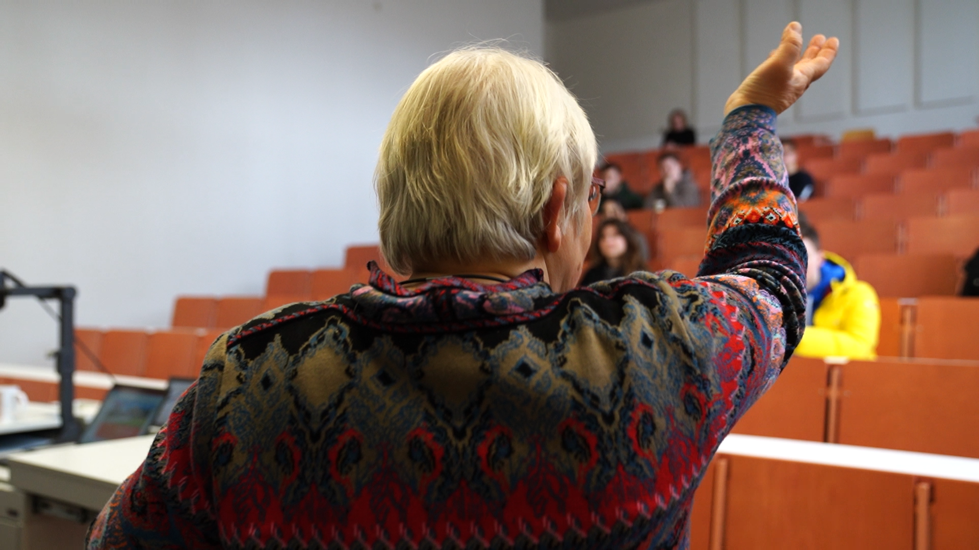 In einem lebhaften Hörsaal der Hochschule Coburg zieht eine Person mit grauem Haar und farbenfroher Kleidung das Publikum in ihren Bann. Ihre erhobene Hand deutet auf orangefarbene Sitzreihen, die mit mehreren aufmerksamen Personen besetzt sind.