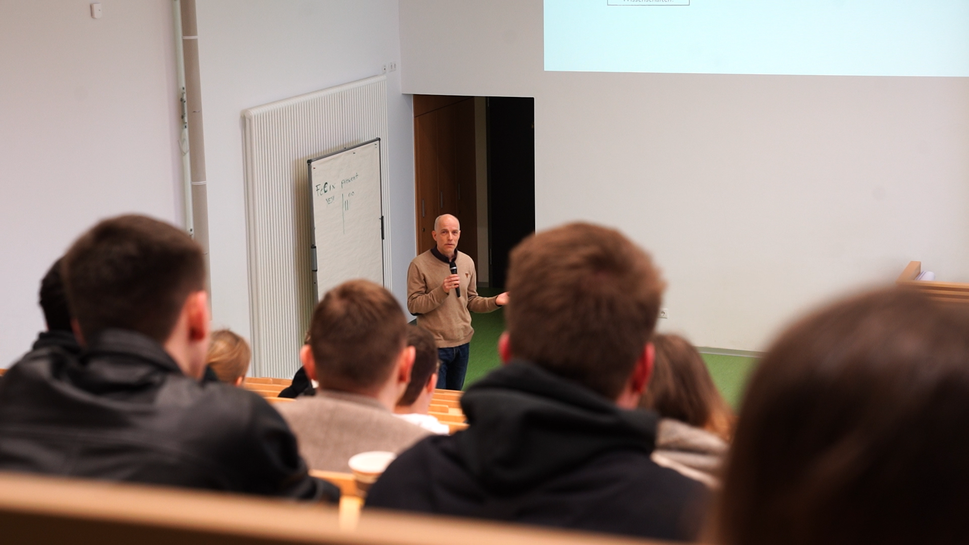 Ein Mann steht vor einem Hörsaal der Hochschule Coburg, hält ein Mikrofon in der Hand und spricht. Hinter ihm ist eine Tafel zu sehen. Die Studierenden sitzen und hören aufmerksam zu.