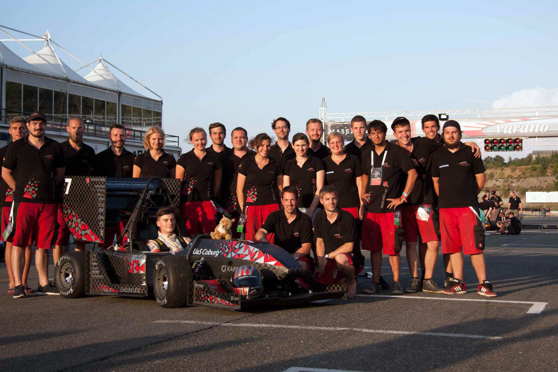 Eine Gruppe von Menschen in passenden schwarzen Shirts und roten Shorts posiert für ein Foto auf der Rennstrecke der Hochschule Coburg neben einem kleinen Rennauto. Im Hintergrund sind Tribünen und ein teilweise sichtbares Schild mit dem Wort „Varano“ zu sehen.