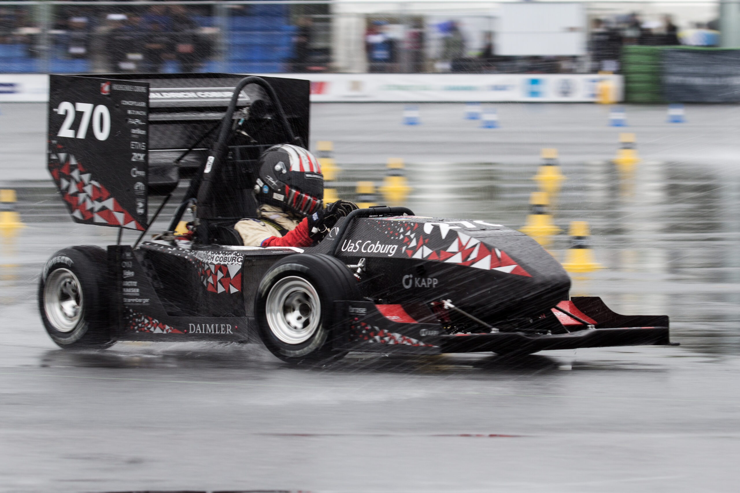 Eine Person fährt einen eleganten, schwarzen Formel-Rennwagen mit der Aufschrift „270“ auf einer nassen Strecke. Das Auto, das stolz die Hochschule Coburg repräsentiert, ist mit rot-weißen geometrischen Mustern und prominenten Sponsoren versehen. Blaue und gelbe Pylonen säumen die Strecke, während Regentropfen den Hintergrund verschwimmen lassen.