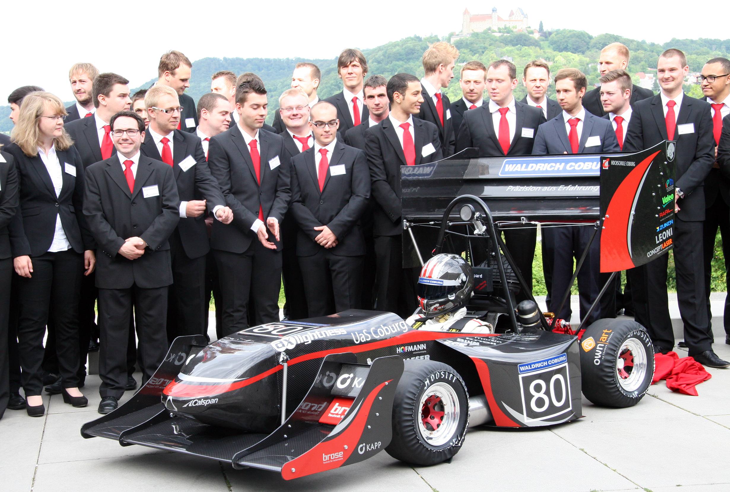 Eine Gruppe von Menschen in formeller Kleidung steht um ein schwarz-rotes Rennauto mit der Nummer 80 und repräsentiert stolz die Hochschule Coburg. Im Hintergrund sind Bäume und ein entferntes Gebäude auf einem Hügel zu sehen, die die Szene für diesen bedeutsamen Anlass bilden.