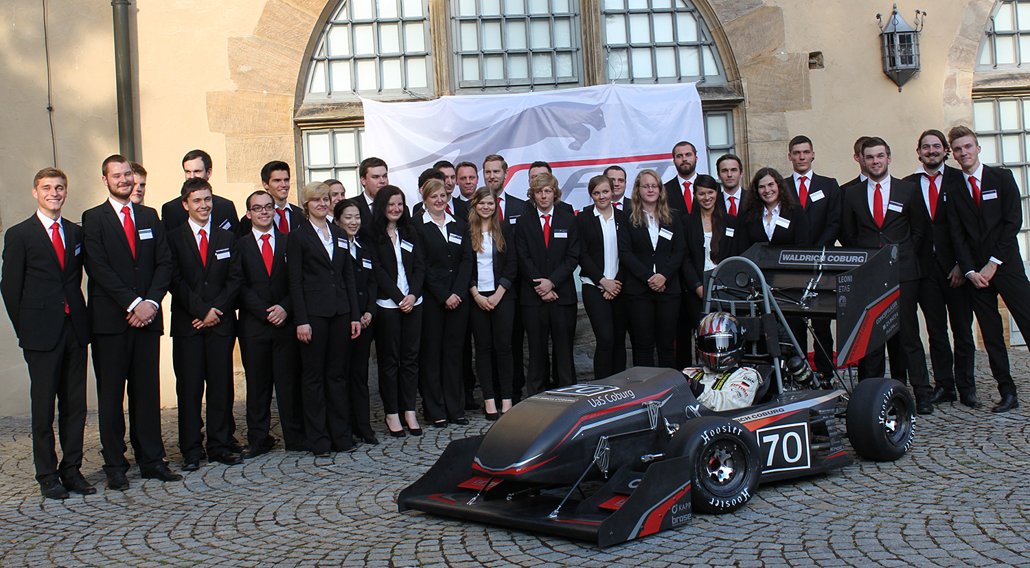 Vor einem Gebäude mit Rundbogenfenstern, das die Hochschule Coburg repräsentiert, steht eine Gruppe gut gekleideter Menschen. Sie versammeln sich um einen kleinen, schnittigen Rennwagen mit der Nummer 70, hinter ihnen hängt ein Banner mit dem Logo der Hochschule.