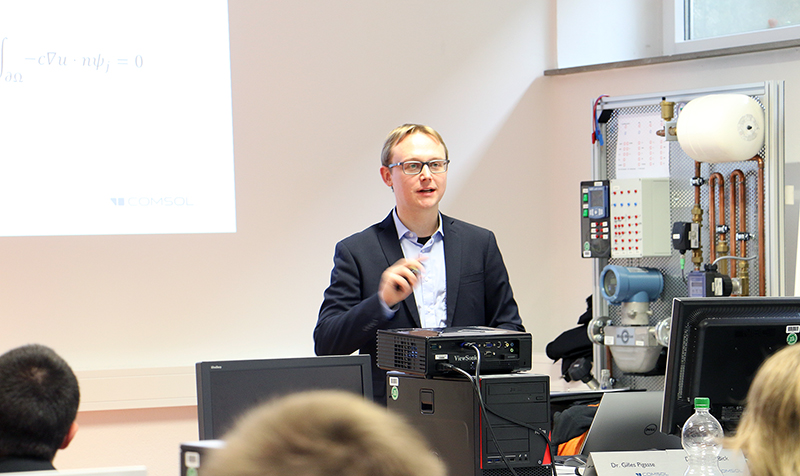 Ein Mann im Anzug spricht vor einem Hörsaal der Hochschule Coburg. Er hält einen Stift in der Hand und steht neben einem elektronischen Gerät, auf dessen Leinwand eine mathematische Gleichung angezeigt wird. Die Teilnehmer sitzen und hören der spannenden Vorlesung aufmerksam zu.