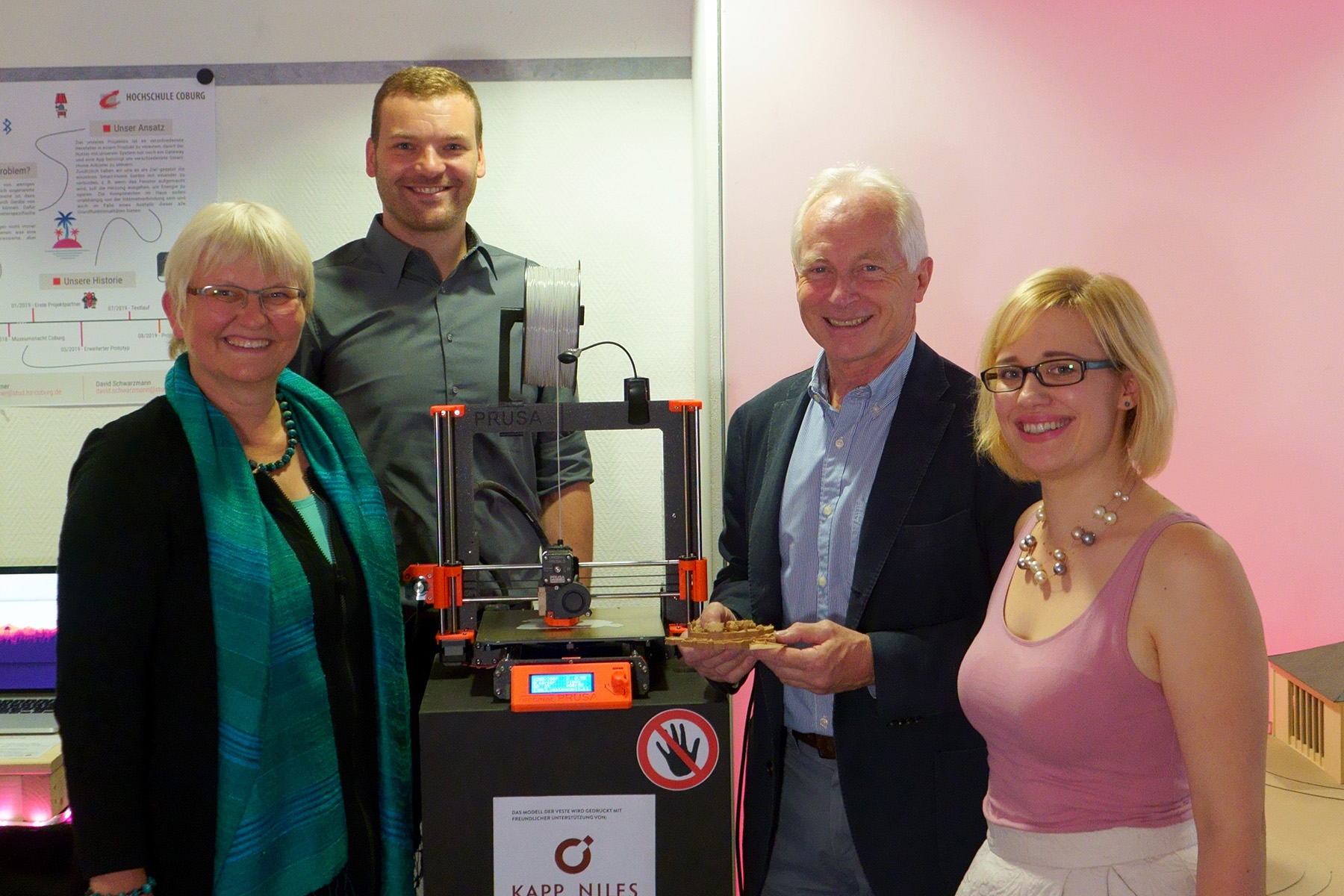 Vier Menschen stehen lächelnd um einen 3D-Drucker in einem Innenbereich der Hochschule Coburg herum. Sie wirken glücklich und engagiert, im Hintergrund sind ein Schild und ein Plakat zu sehen. Die Atmosphäre ist freundlich und professionell.