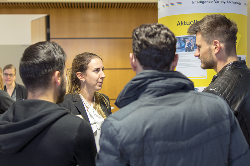 Bei der Veranstaltung der Hochschule Coburg unterhält sich eine Frau im Business-Anzug mit vier Männern, die der Kamera den Rücken zukehren. Sie stehen vor einem Display mit teilweise lesbarem Text, was eine Konferenzatmosphäre in Innenräumen suggeriert.