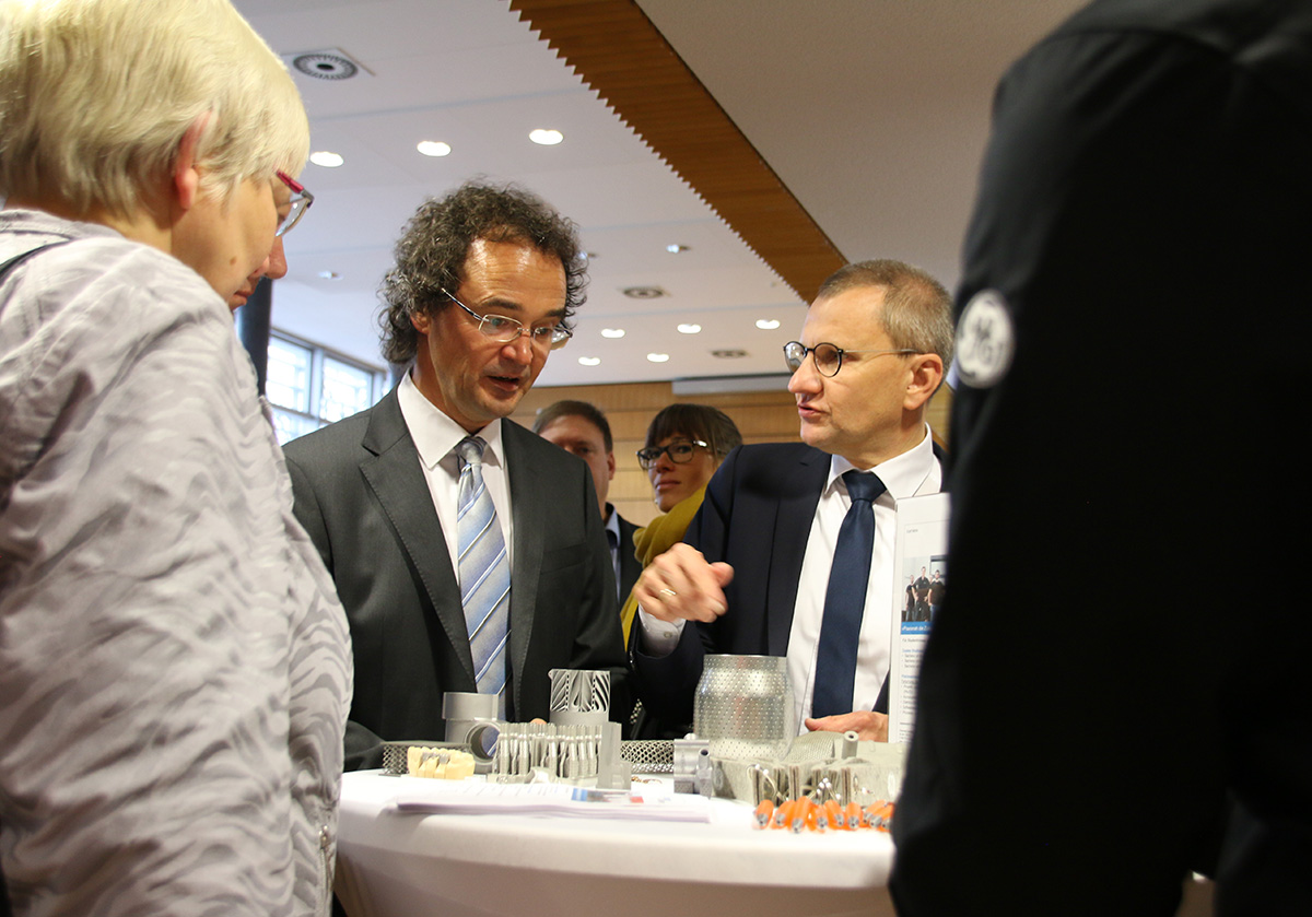 Eine Gruppe von Fachleuten in Business-Kleidung versammelt sich um einen Tisch in einem Gebäude, das wie ein Konferenzraum der Hochschule Coburg aussieht. Im Mittelpunkt stehen zwei Männer, die sich unterhalten, während die anderen aufmerksam zuhören, umgeben von verschiedenen Gegenständen, die auf kollaborative Innovation hindeuten.