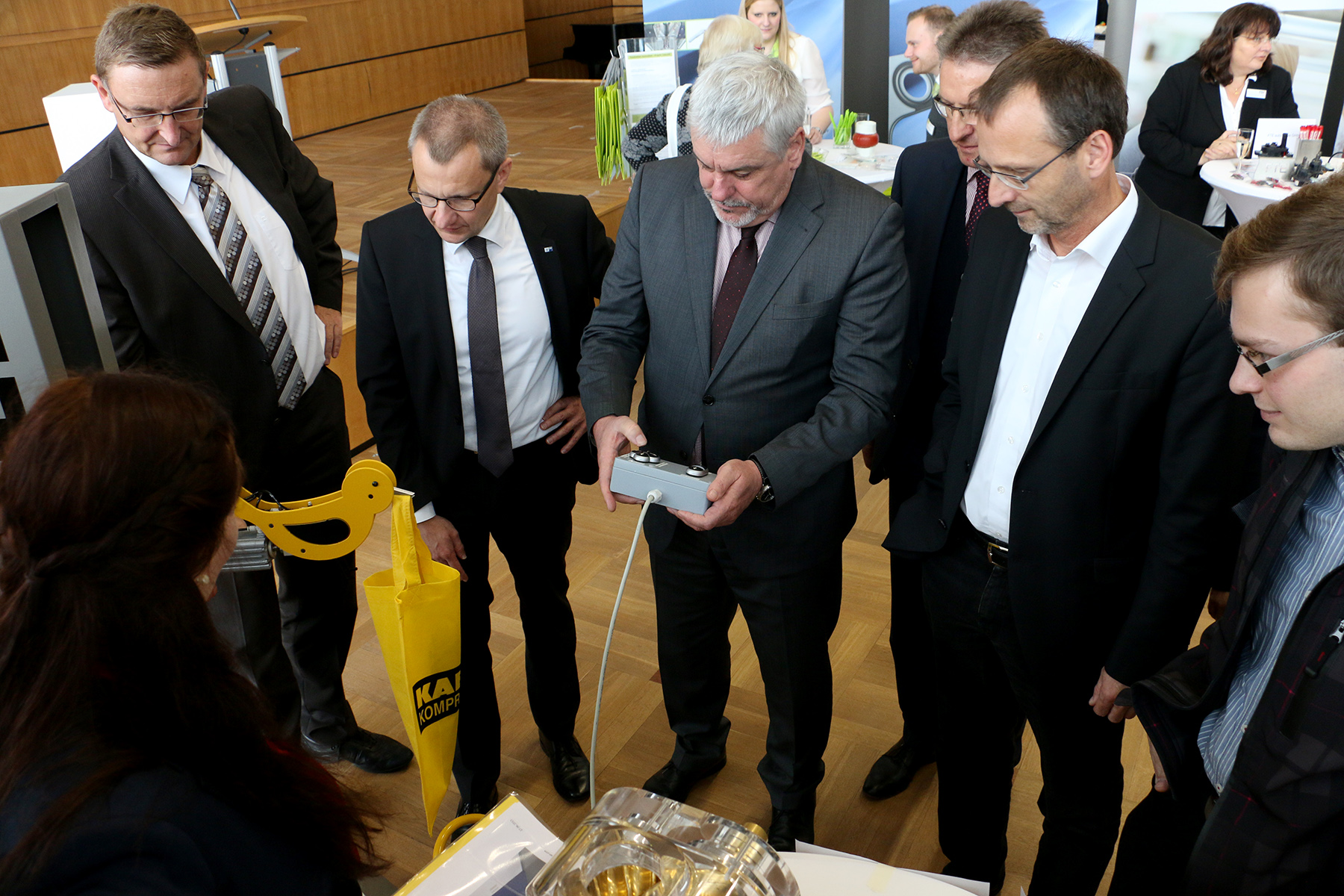 Eine Gruppe von sieben Personen in Business-Kleidung steht um einen Tisch herum und beobachtet aufmerksam einen Mann im grauen Anzug, der ein Gerät hält. Die Umgebung erinnert an den formellen Besprechungsraum der Hochschule Coburg und besteht aus Ausstellungsstücken auf Tischen.