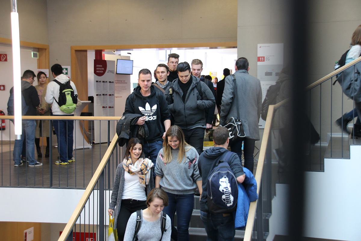 Eine Gruppe von Menschen, darunter Männer und Frauen, geht in einem belebten Innenbereich der Hochschule Coburg eine Treppe auf und ab. Einige tragen Rucksäcke und legere Kleidung wie Jacken und Sweatshirts. Der Bereich ähnelt einem öffentlichen Raum, möglicherweise innerhalb der lebhaften Campusumgebung.