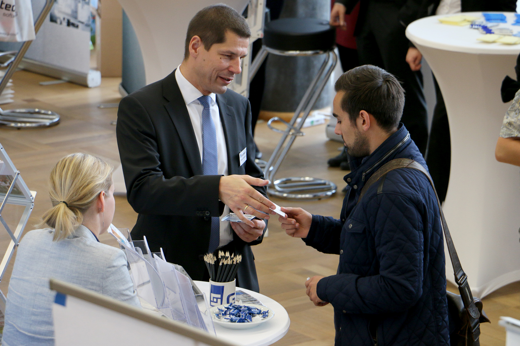 Bei einer Networking-Veranstaltung überreicht ein Mann im Anzug einem anderen im blauen Jackett seine Visitenkarte und erwähnt ihr gemeinsames Interesse an der Hochschule Coburg. Daneben sitzt eine blonde Frau mit Broschüren und Stiften an einem Tisch. Im Raum herrscht reges Treiben, da sich im Hintergrund an hohen Tischen Menschen unterhalten.