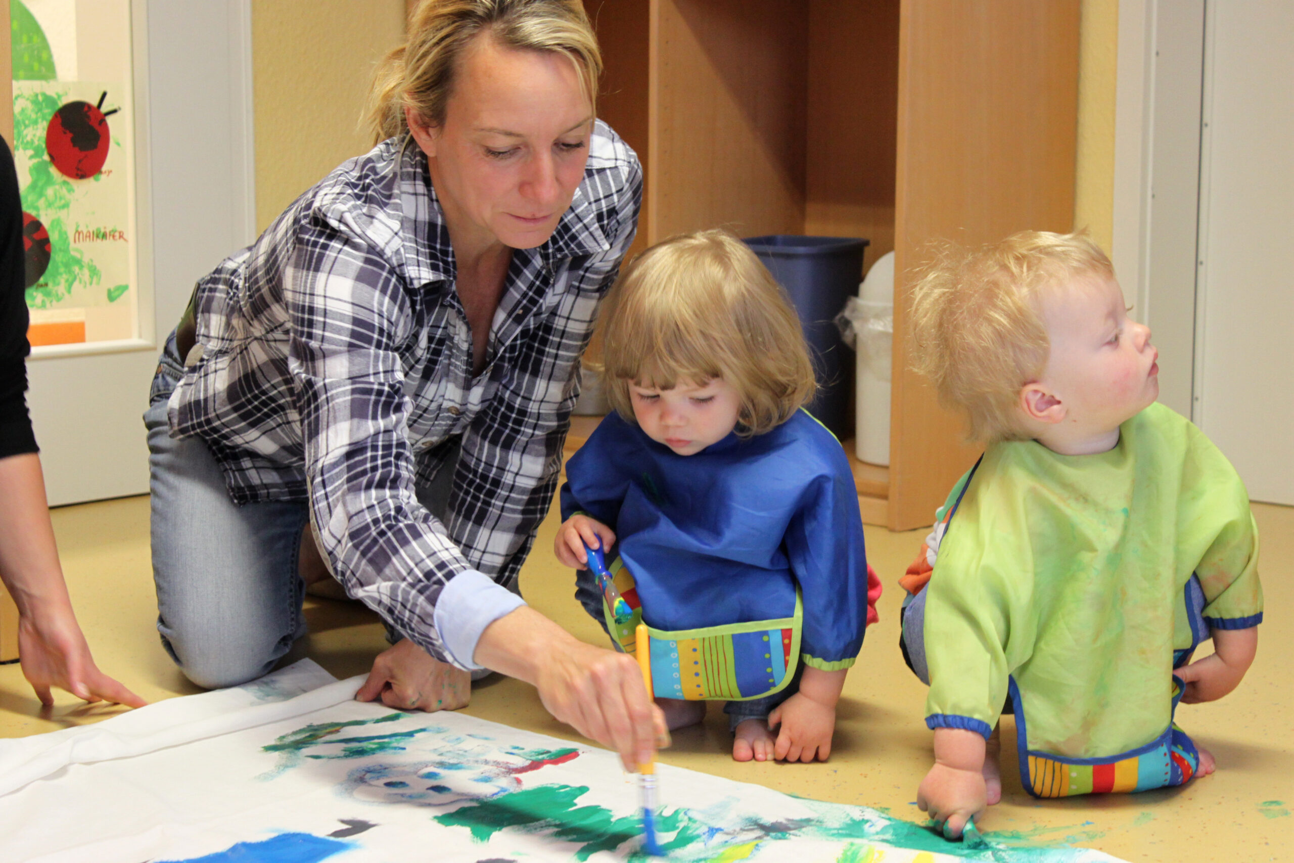 Ein Erwachsener und zwei kleine Kinder sind an der Hochschule Coburg bei einer Malaktivität dabei. Die Kinder sitzen in Schutzkitteln mit Pinseln auf dem Boden, während der Erwachsene ihnen hilft, ihre künstlerischen Ausdrucksformen auf einem großen weißen Blatt zu lenken. Farbenfrohe Kunst schmückt den Hintergrund.