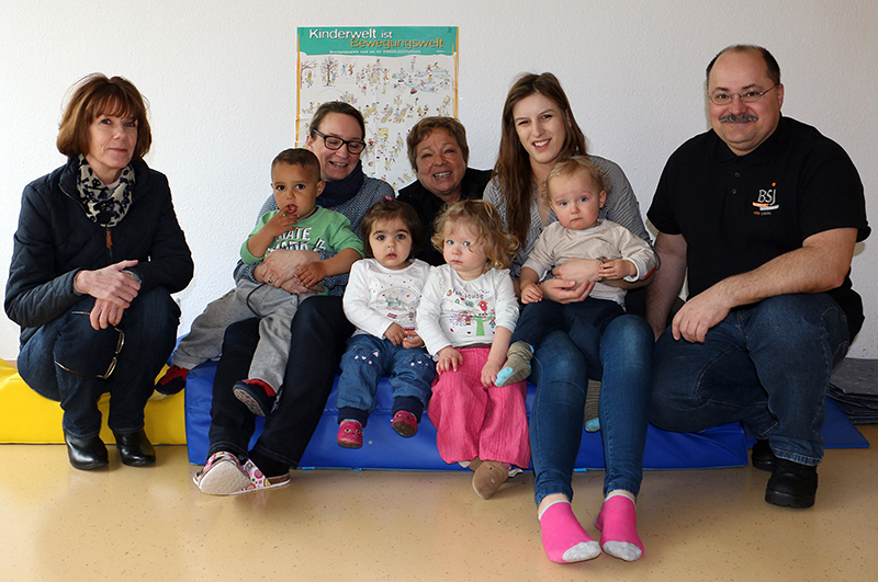 Eine Gruppe Erwachsener und Kinder sitzt drinnen auf einer bunten Matte zusammen, vermutlich bei einer Veranstaltung der Hochschule Coburg. Drei Frauen, zwei Männer und vier kleine Kinder sind überall lächelnd zu sehen. Es herrscht eine warme und einladende Atmosphäre, die durch das farbenfrohe Poster an der Wand hinter ihnen noch verstärkt wird.