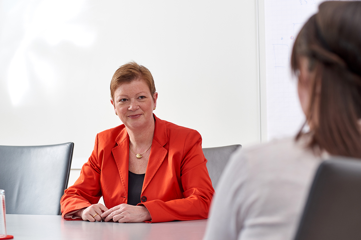 Eine Frau im orangefarbenen Blazer sitzt an einem Konferenztisch und hört einer anderen Person aufmerksam zu, die mit dem Rücken zur Kamera steht. Sie befinden sich in einem hellen, modernen Besprechungsraum der Hochschule Coburg, im Hintergrund ein Whiteboard.