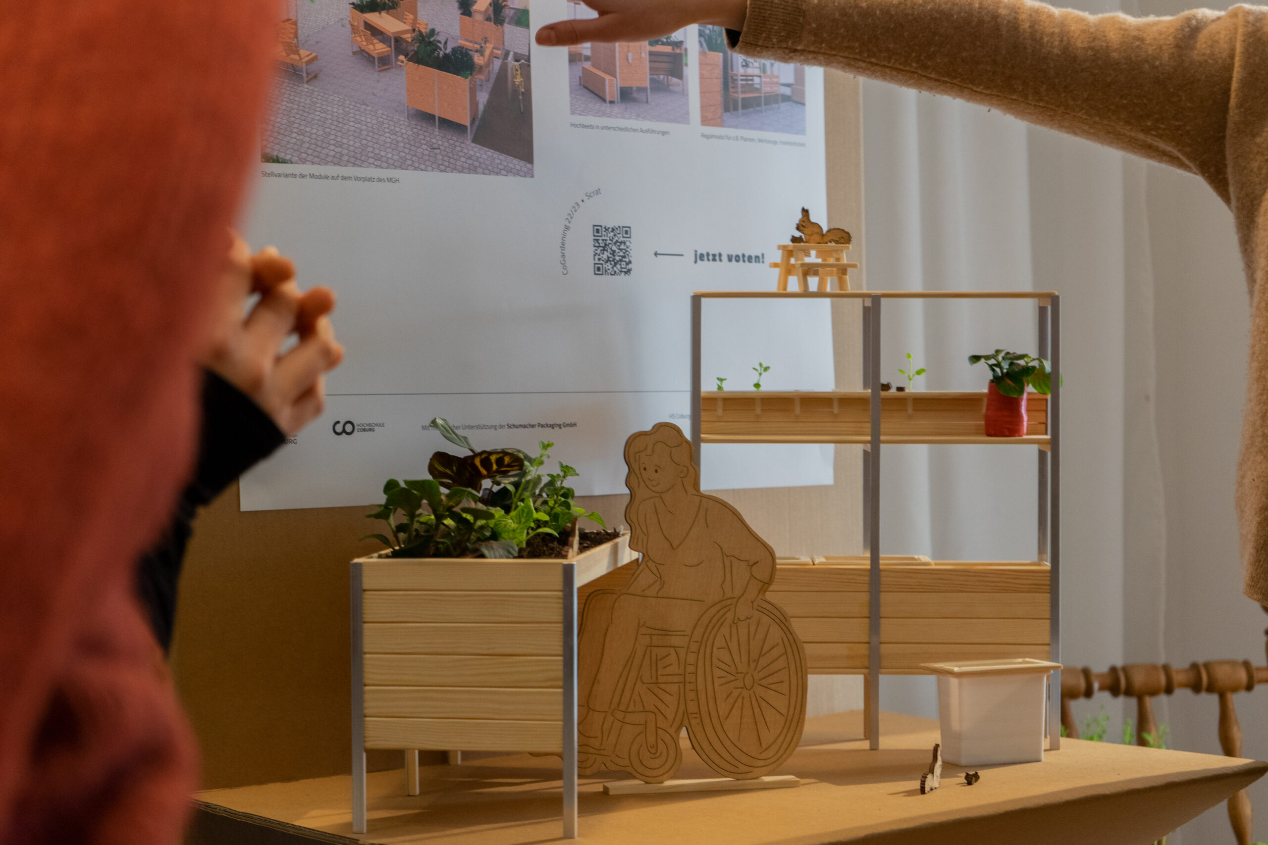 Eine Person deutet auf ein kleines Modell eines barrierefreien Gartens an der Hochschule Coburg. Das Modell besteht aus einem erhöhten Pflanzkasten aus Holz mit einer Figur im Rollstuhl, Regalen mit kleinen Pflanzen und einem Poster im Hintergrund, auf dem entsprechende Bilder zu sehen sind.