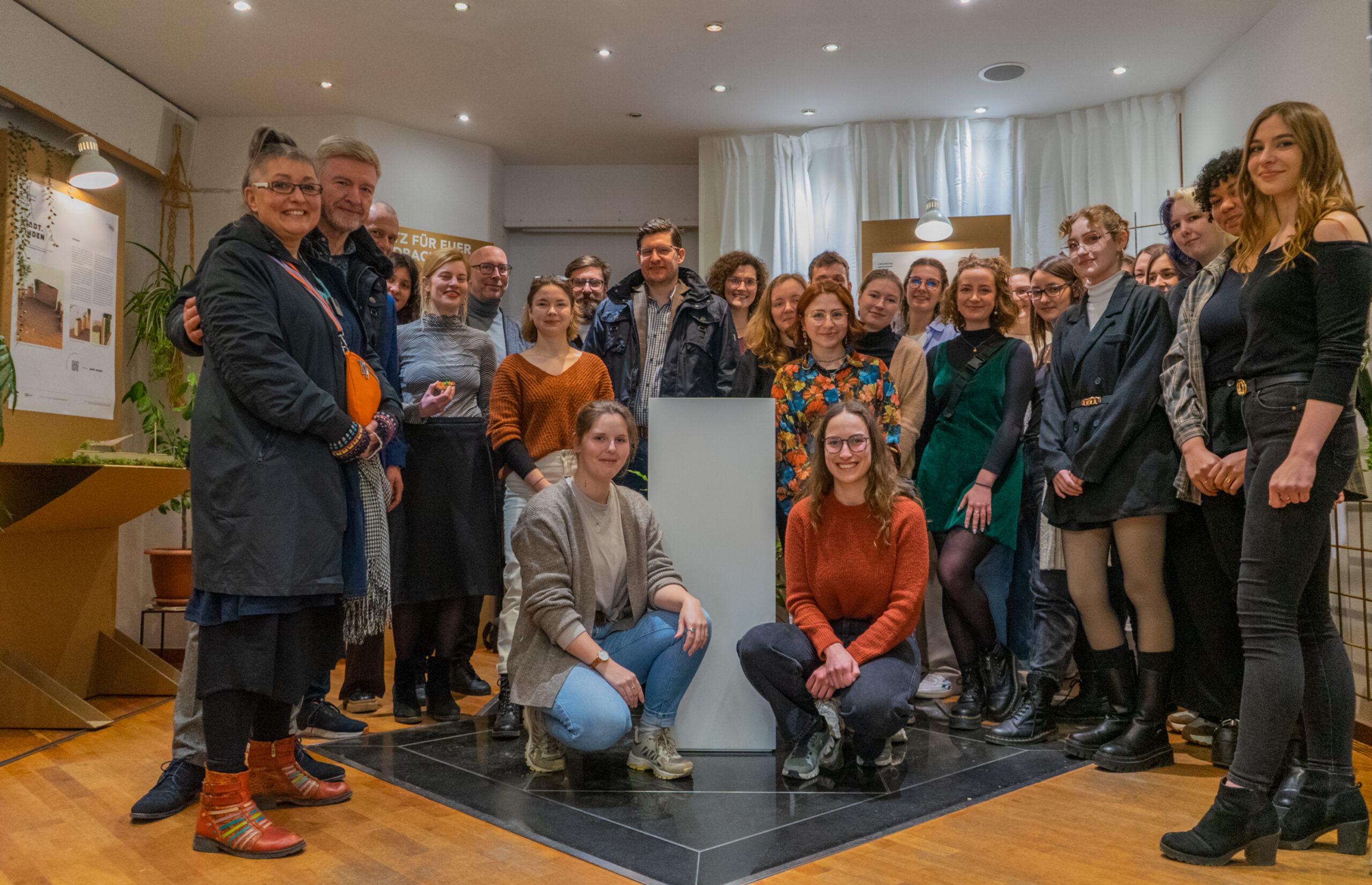 Eine Gruppe von Menschen posiert gemeinsam in einem gut beleuchteten Raum der Hochschule Coburg mit Holzboden und weißen Wänden. Sie stehen um einen weißen Sockel herum und lächeln in die Kamera. Verschiedene Pflanzen und Kunstwerke schmücken den Hintergrund und verleihen dem akademischen Ambiente Charme.