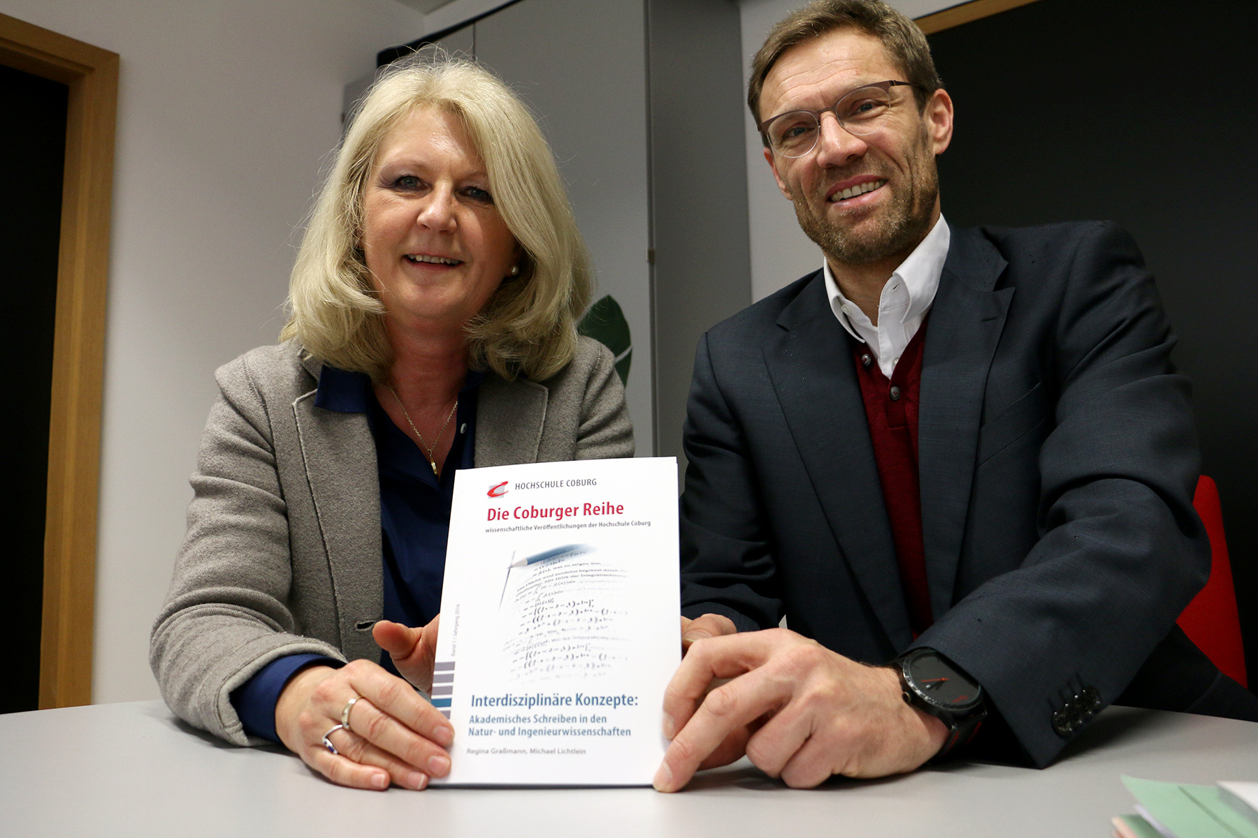 Eine Frau und ein Mann sitzen lächelnd an einem Tisch und halten ein Buch mit dem Titel „Die Coburger Reiche“ in den Händen. Darin werden interdisziplinäre Konzepte der Hochschule Coburg sowie der Universitäten Augsburg und Ingolstadt vorgestellt. Sie sind in Business-Kleidung gekleidet und strahlen Professionalität und Teamgeist aus.