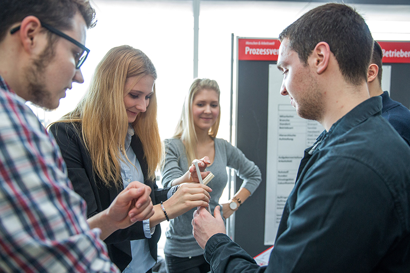 Eine Gruppe von Menschen an der Hochschule Coburg diskutiert und untersucht in einem hellen Raum ein Objekt. Sie sind leger gekleidet und schaffen so eine kollaborative und konzentrierte Atmosphäre. Im Hintergrund unterstützen informative Poster oder Schautafeln ihre Erkundung.