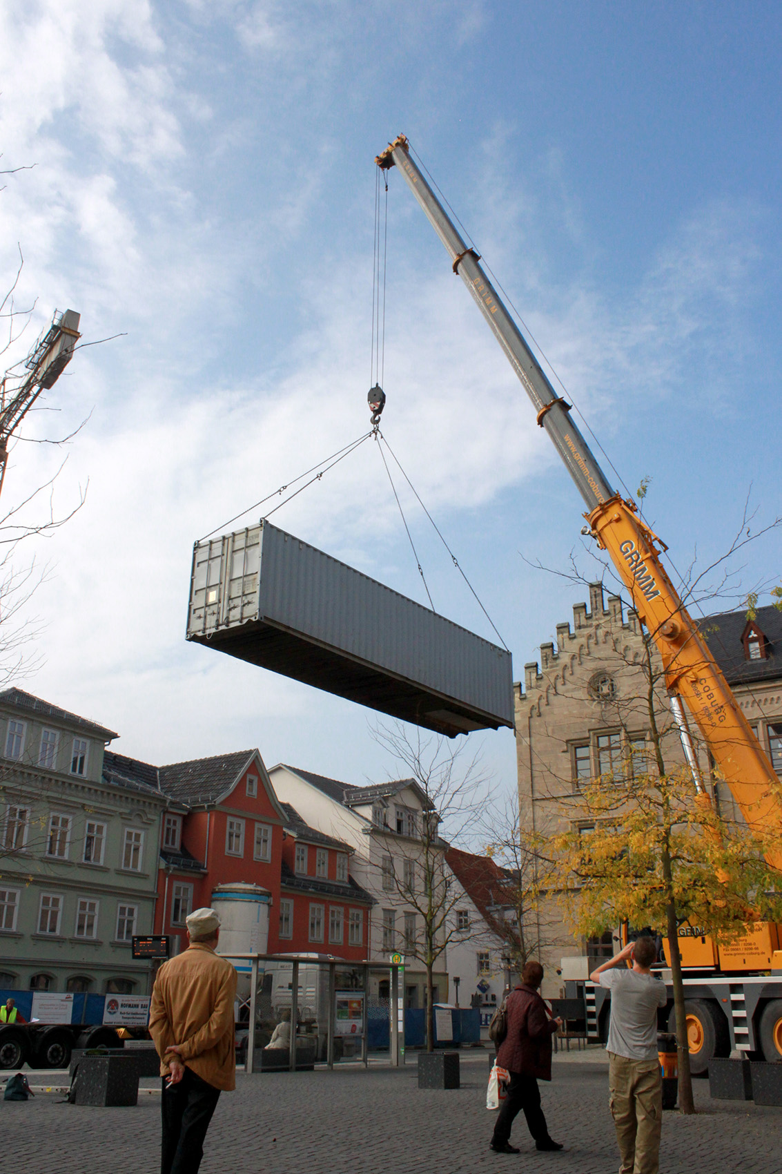 Ein Kran hebt einen großen Schiffscontainer auf einen Stadtplatz in der Nähe der Hochschule Coburg. Menschen beobachten die Aktion, im Hintergrund stehen historische Gebäude mit Satteldächern unter einem klaren blauen Himmel.