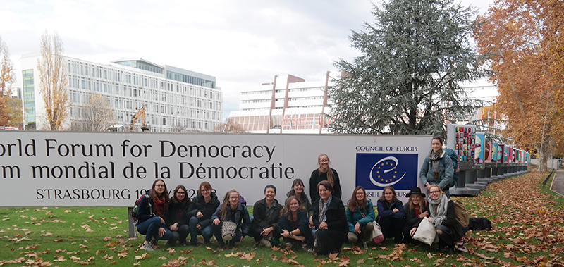 Eine Gruppe von Menschen der Hochschule Coburg sitzt und kniet im Gras vor einem großen Schild mit der Aufschrift „Weltforum für Demokratie, Europarat“. Gebäude und Herbstbäume bilden eine malerische Kulisse.