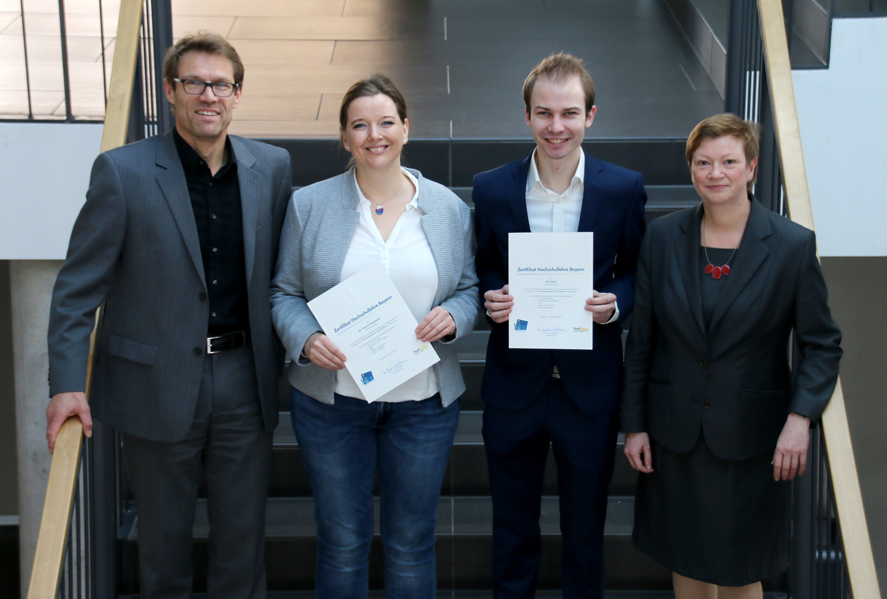 Vier Personen stehen auf einer Treppe im Innenbereich der Hochschule Coburg. Zwei in der Mitte halten lächelnd Zertifikate in den Händen, während zwei weitere auf beiden Seiten stehen. Sie sind in Business-Kleidung gekleidet und posieren für ein Gruppenfoto.