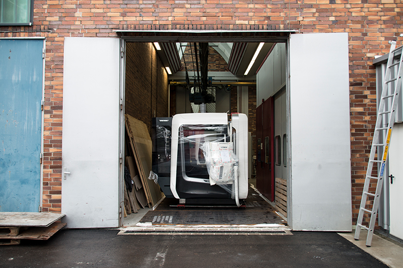 Eine große Maschine für die Hochschule Coburg wird durch ein weit geöffnetes Tor in eine gemauerte Industriehalle transportiert. Verpackungsmaterial ist auf der Maschine zu sehen, Paletten und Leitern stehen neben dem Eingang und erleichtern die Installation.