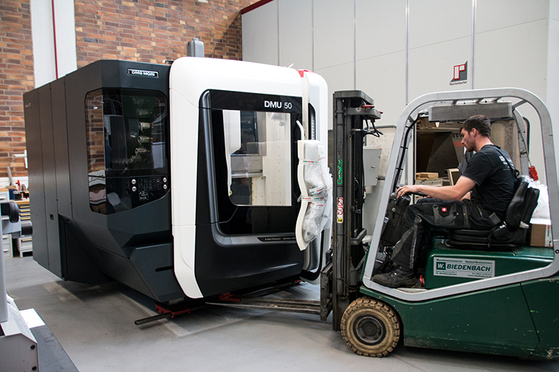 Ein Arbeiter bedient einen grünen Gabelstapler, um eine große DMU 50-Maschine in einem Industriegebäude der Hochschule Coburg zu positionieren. Im Hintergrund sind eine Backsteinmauer und moderne Geräte zu sehen, die die fortschrittlichen Einrichtungen der Hochschule hervorheben.