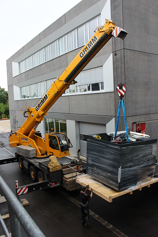 Vor dem modernen, mehrstöckigen Gebäude der Hochschule Coburg hebt ein großer gelber Kran eine Palette an, die mit schwarzem Material und blauen Gurten umwickelt ist. Eine Person steht daneben und leitet den Vorgang auf der gepflasterten Fläche.