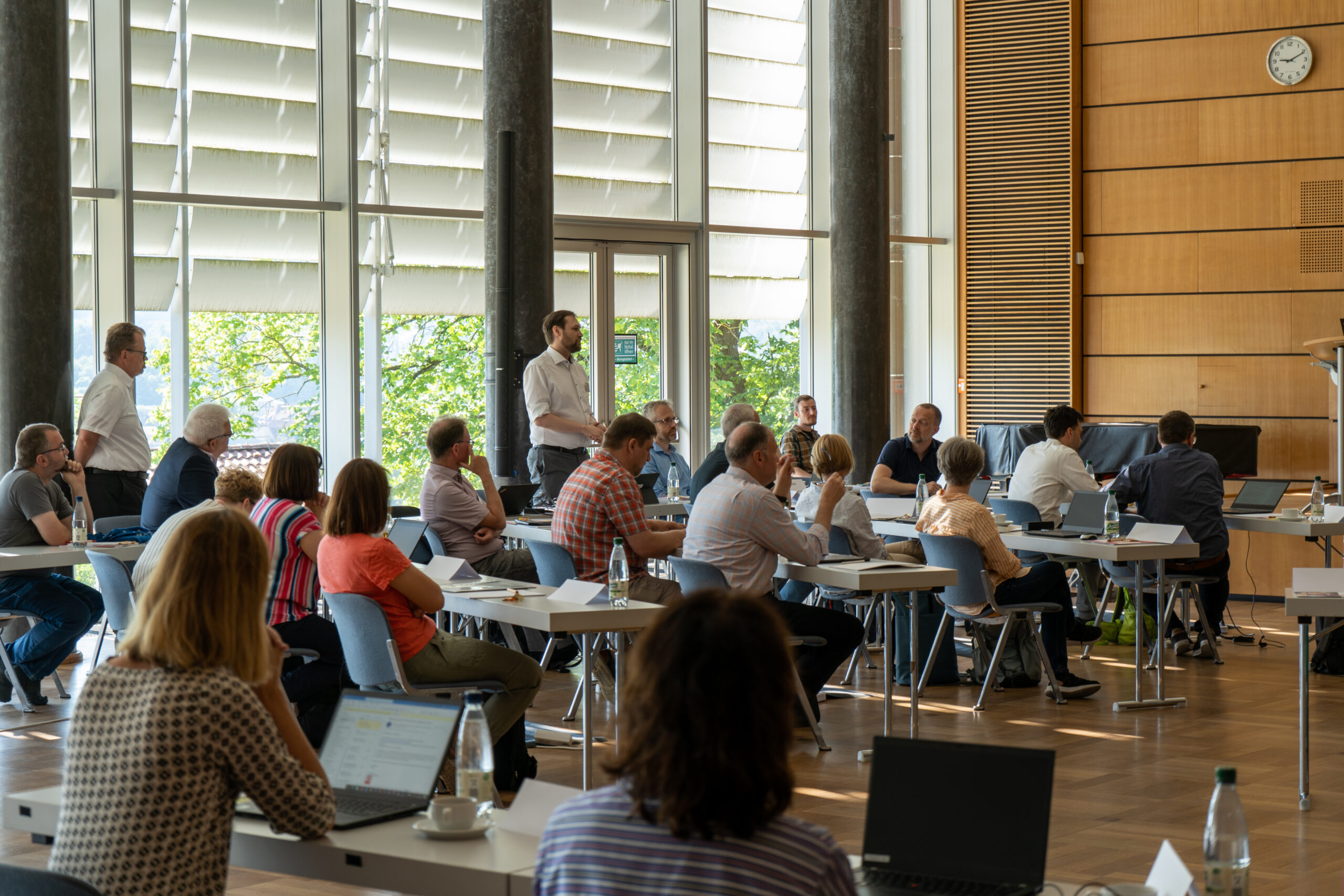 Eine Gruppe von Menschen sitzt in einem geräumigen, sonnendurchfluteten Raum mit großen Fenstern. Einige arbeiten an Laptops, während andere einem links stehenden Redner zuhören. Diese lebhafte Szene an der Hochschule Coburg lässt an eine Workshop- oder Konferenzatmosphäre denken.