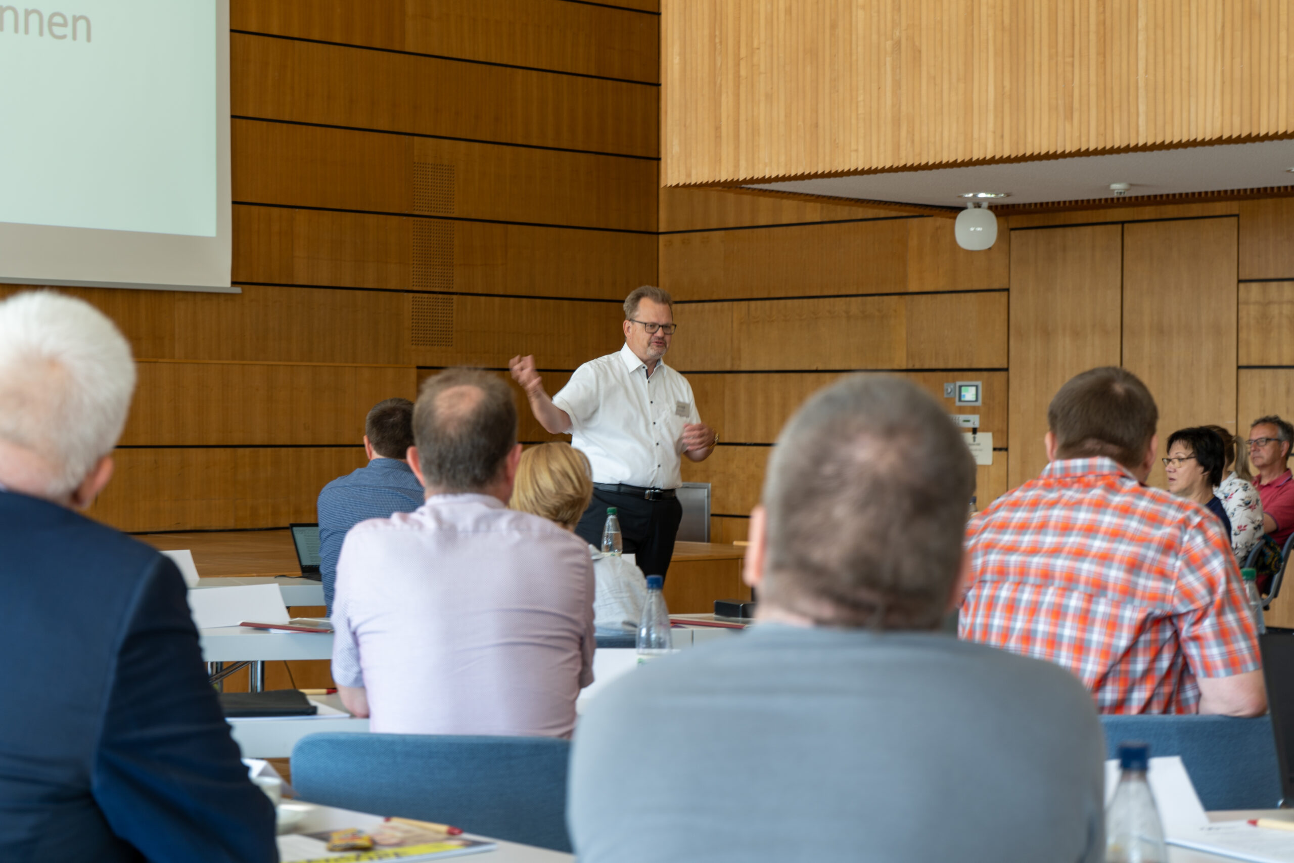 Ein Mann steht vor einem Konferenzraum der Hochschule Coburg und gestikuliert, während er zu einem sitzenden Publikum spricht. Der Raum hat Holzwände und die Teilnehmer sind aufmerksam und sitzen mit Unterlagen und Wasserflaschen an Tischen.