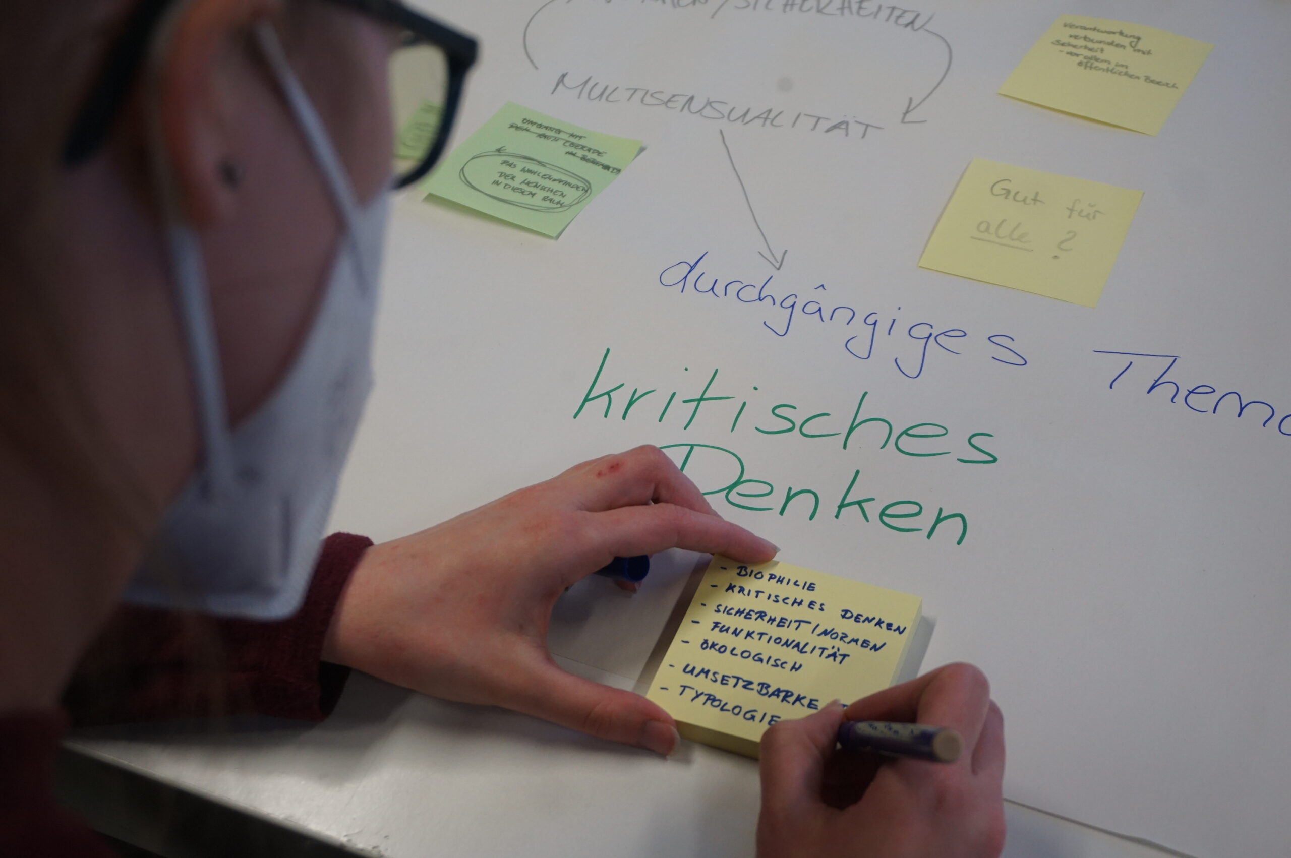 A person wearing glasses and a mask writes on a sticky note at a table. Among the scattered handwritten notes is the phrase "kritisches Denken" in green, surrounded by colorful sticky notes. This vibrant scene captures the spirit of inquiry often found at Hochschule Coburg.