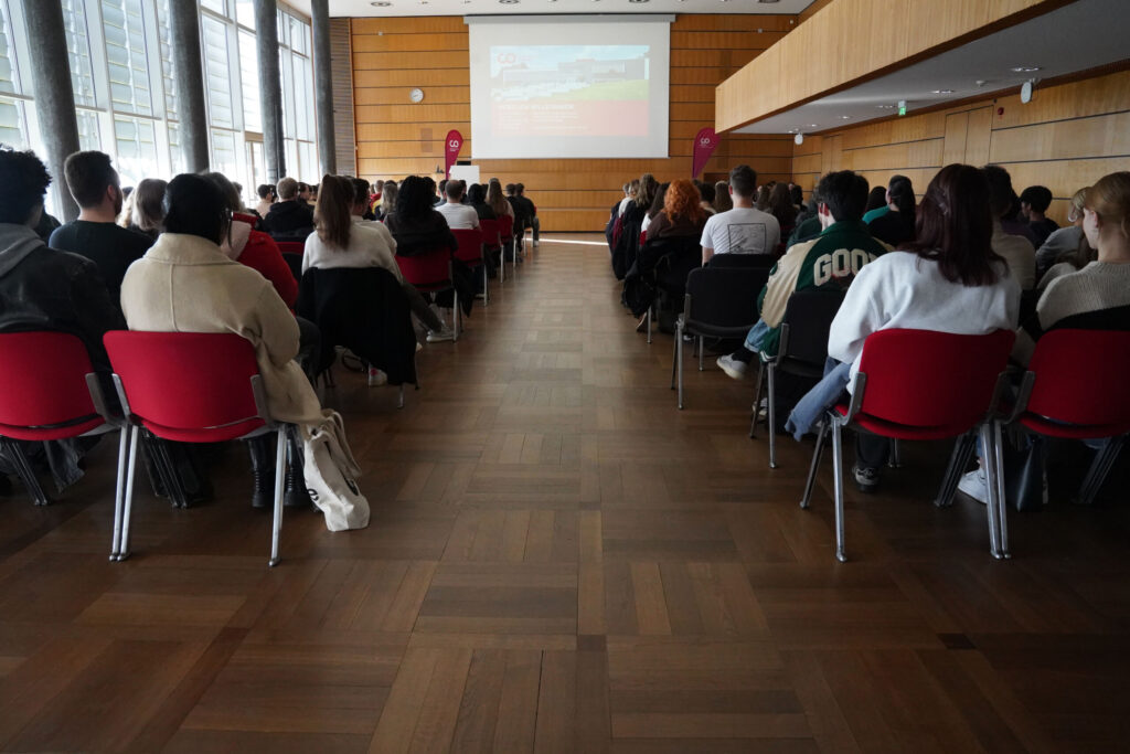 Eine bunt gemischte Gruppe sitzt in Reihen roter Stühle in einem großen, modernen Konferenzraum mit Holzvertäfelung an der Hochschule Coburg. Sie blicken auf einen Bildschirm, auf dem vorne im Raum eine Präsentation läuft.