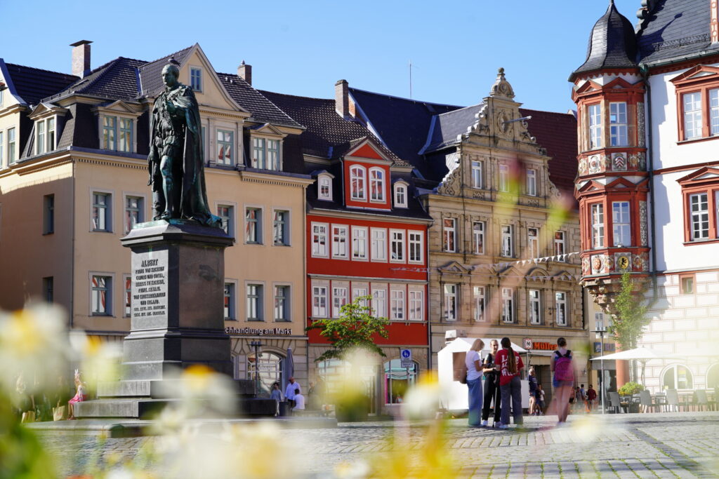 Auf einem belebten Stadtplatz in der Nähe der Hochschule Coburg steht eine Bronzestatue auf einem Sockel, umgeben von historischen Gebäuden mit farbenfrohen Fassaden. Blumen im Vordergrund sorgen für Lebendigkeit, während sich eine Gruppe von Menschen versammelt und die angenehme Atmosphäre unter einem klaren blauen Himmel genießt.