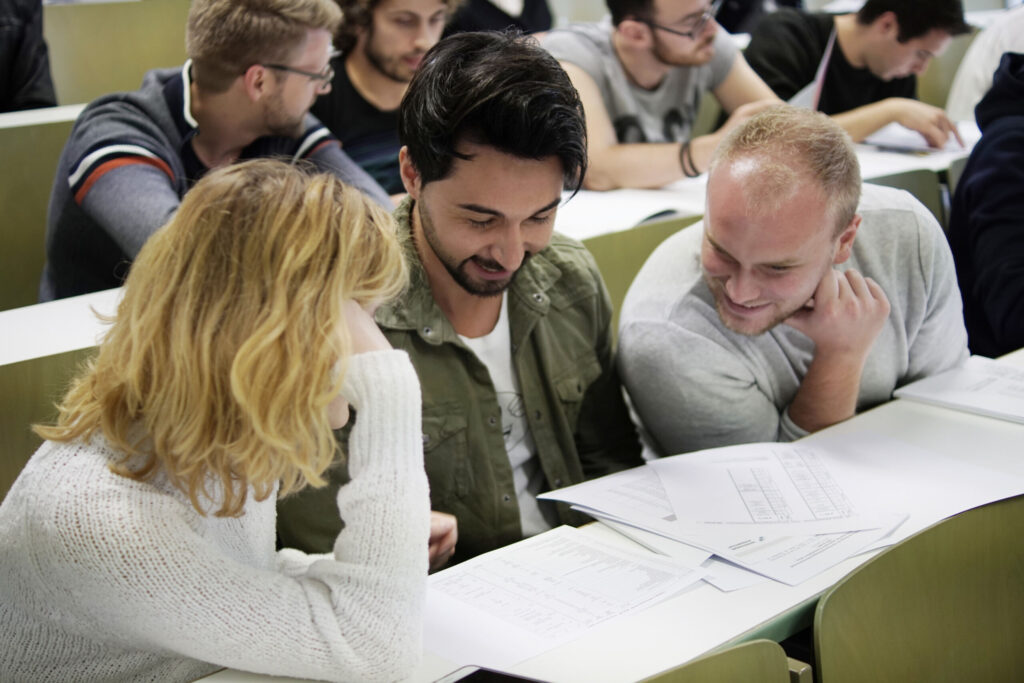 Drei Studierende sitzen an einem Schreibtisch in einem Hörsaal der Hochschule Coburg und arbeiten gemeinsam an einem Dokument. Sie lächeln und wirken engagiert, im Hintergrund sind andere Studierende zu sehen.