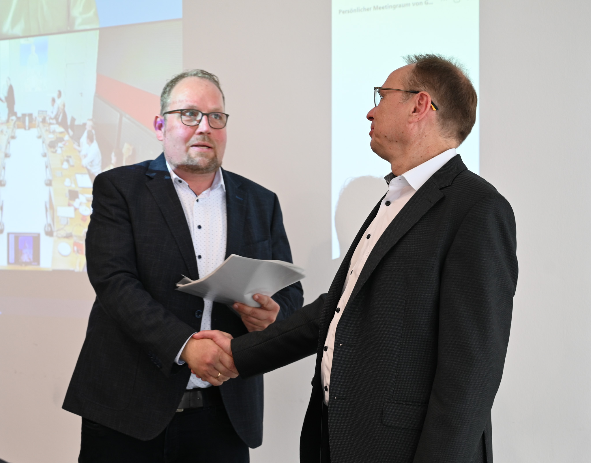 Two men in suits shake hands in an office setting, a digital presentation featuring Hochschule Coburg in the background. One man holds papers, and they appear to be engaged in a formal business meeting.