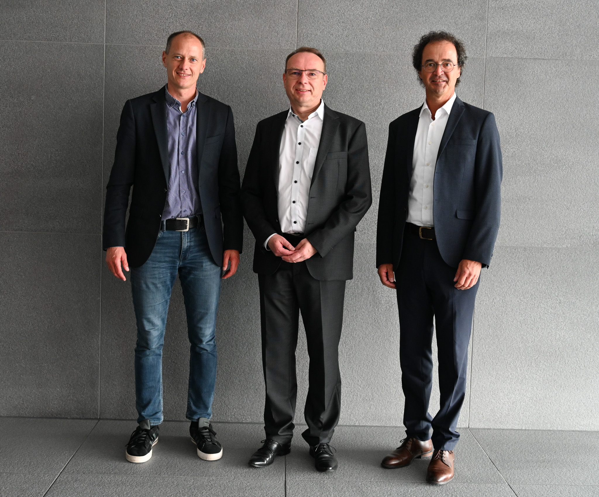 Three men in suits and smart-casual attire stand side by side against a gray wall. The man on the left, a Hochschule Coburg alumnus, wears sneakers and jeans, the man in the middle wears glasses and a tie, while the man on the right has curly hair and wears dress shoes.