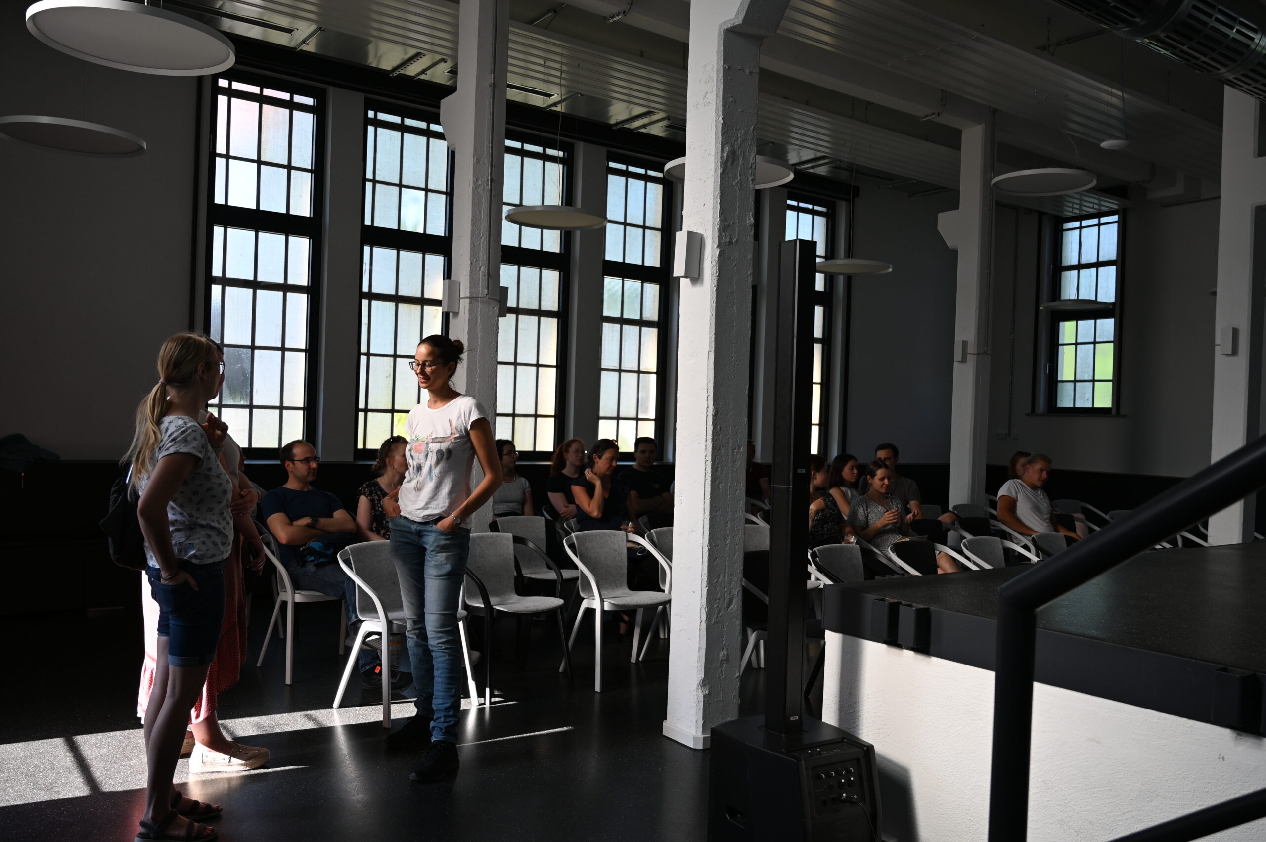 Two women stand and talk in a well-lit room with large windows at Hochschule Coburg. Several people are seated on chairs, listening attentively, suggesting a casual meeting or lecture setting.