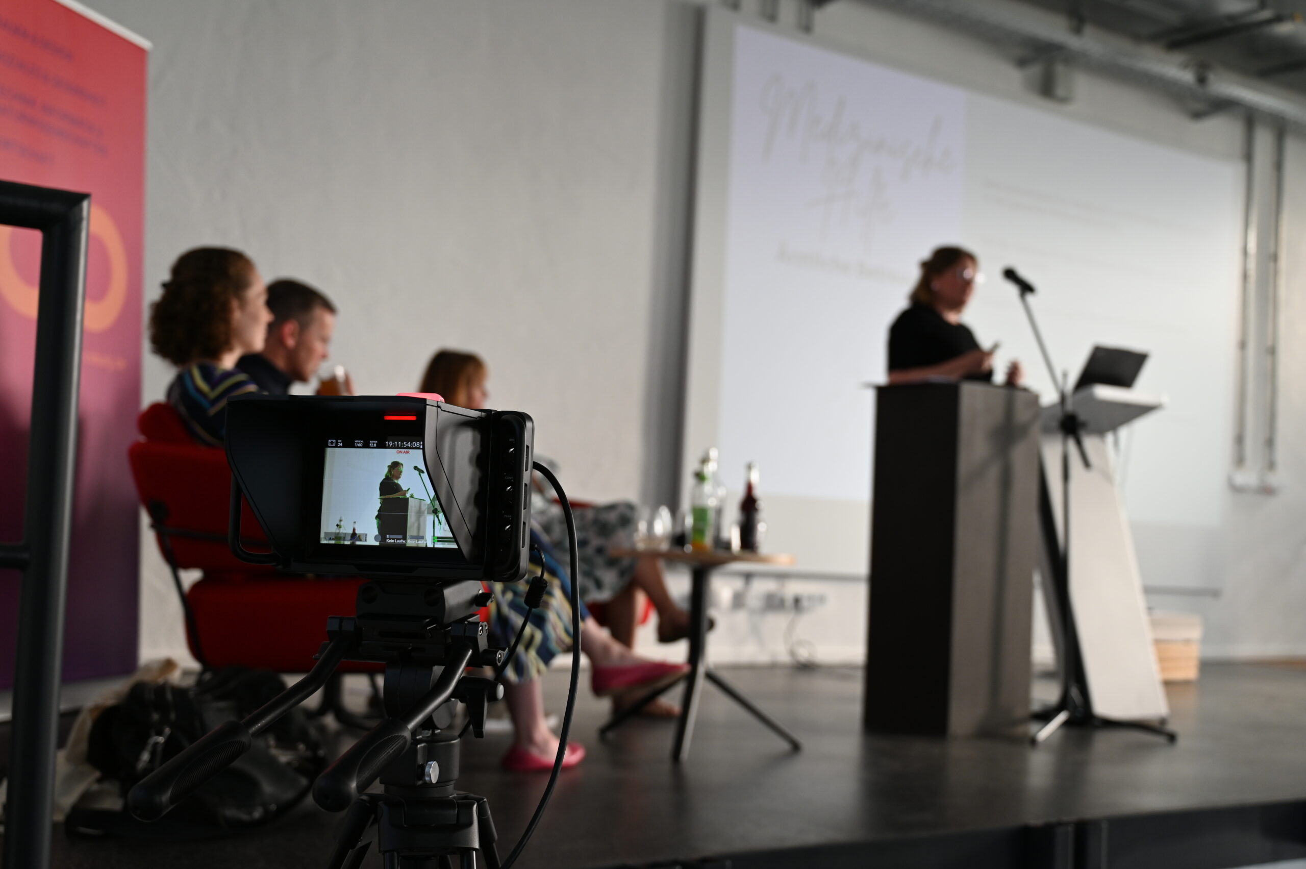 During a presentation at Hochschule Coburg, a panel of four sits on stage as a speaker stands at the podium, microphone in hand. A camera in the foreground records the event, capturing the speaker’s every word. Nearby, bottles and a laptop are stationed on a table.