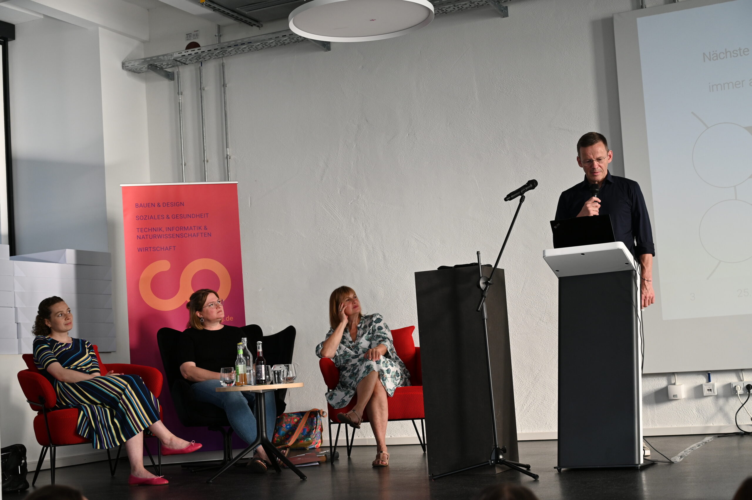 A man speaks at a podium in a conference room at Hochschule Coburg, while three people sit in chairs looking on. A banner with text and a logo is on the left, and a projected screen is partially visible on the right.