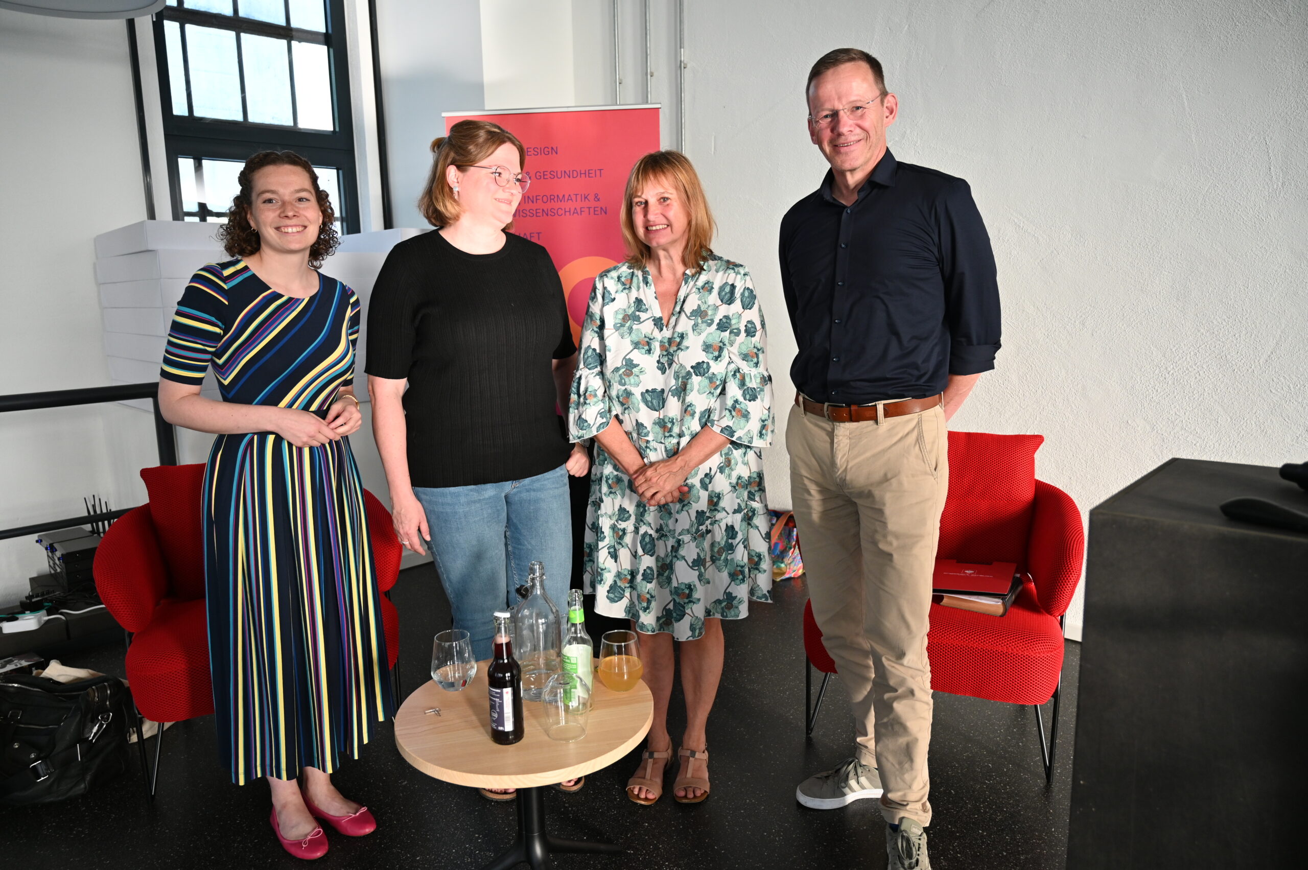 Four people stand together in a room with two red chairs and a small table with drinks. The group, affiliated with Hochschule Coburg, includes three women and one man, all smiling. They are casually dressed, with a poster and windows in the background.